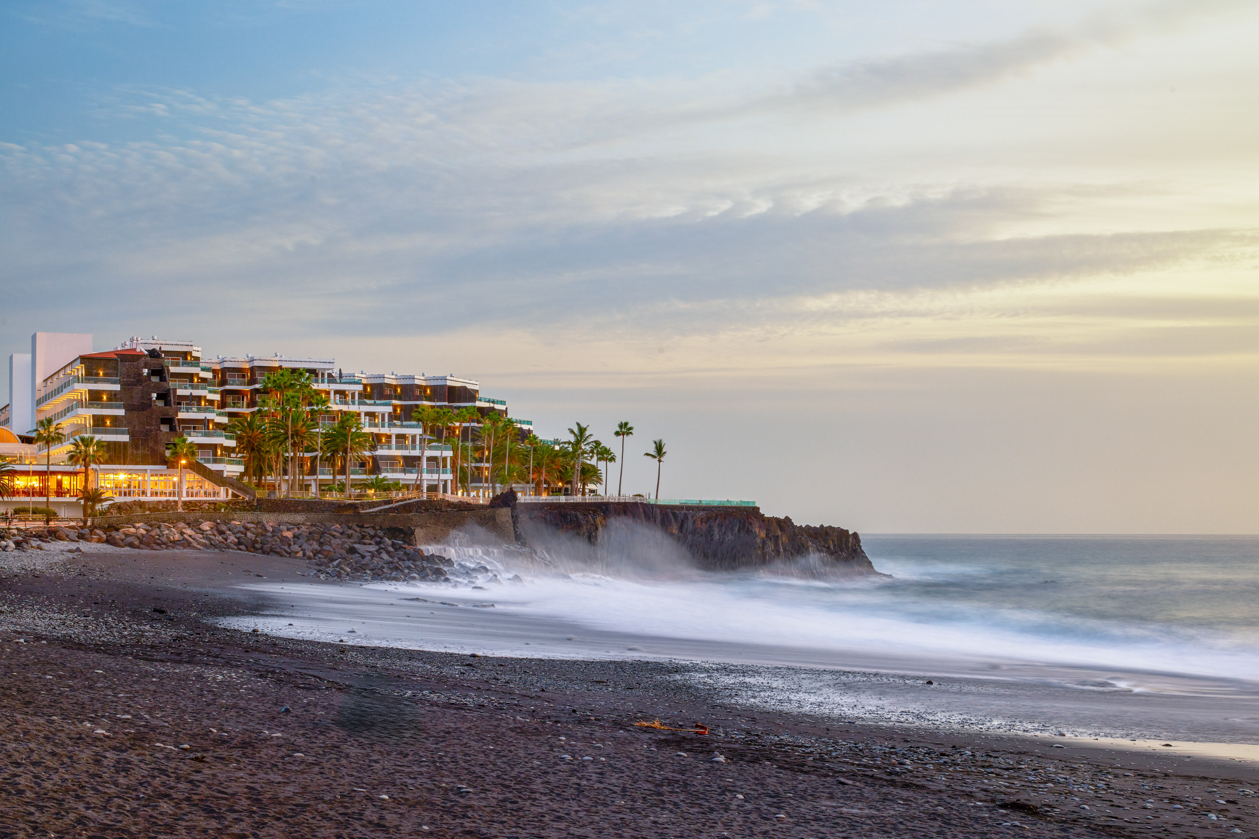 Coastal hotel on cliff, black sand beach, palm trees, and ocean waves at dusk.