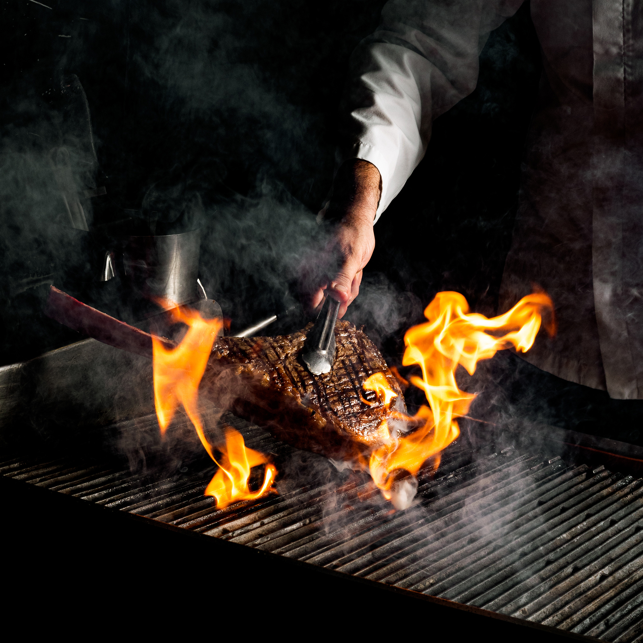 Chef grilling steak with tongs over open flames and smoke.