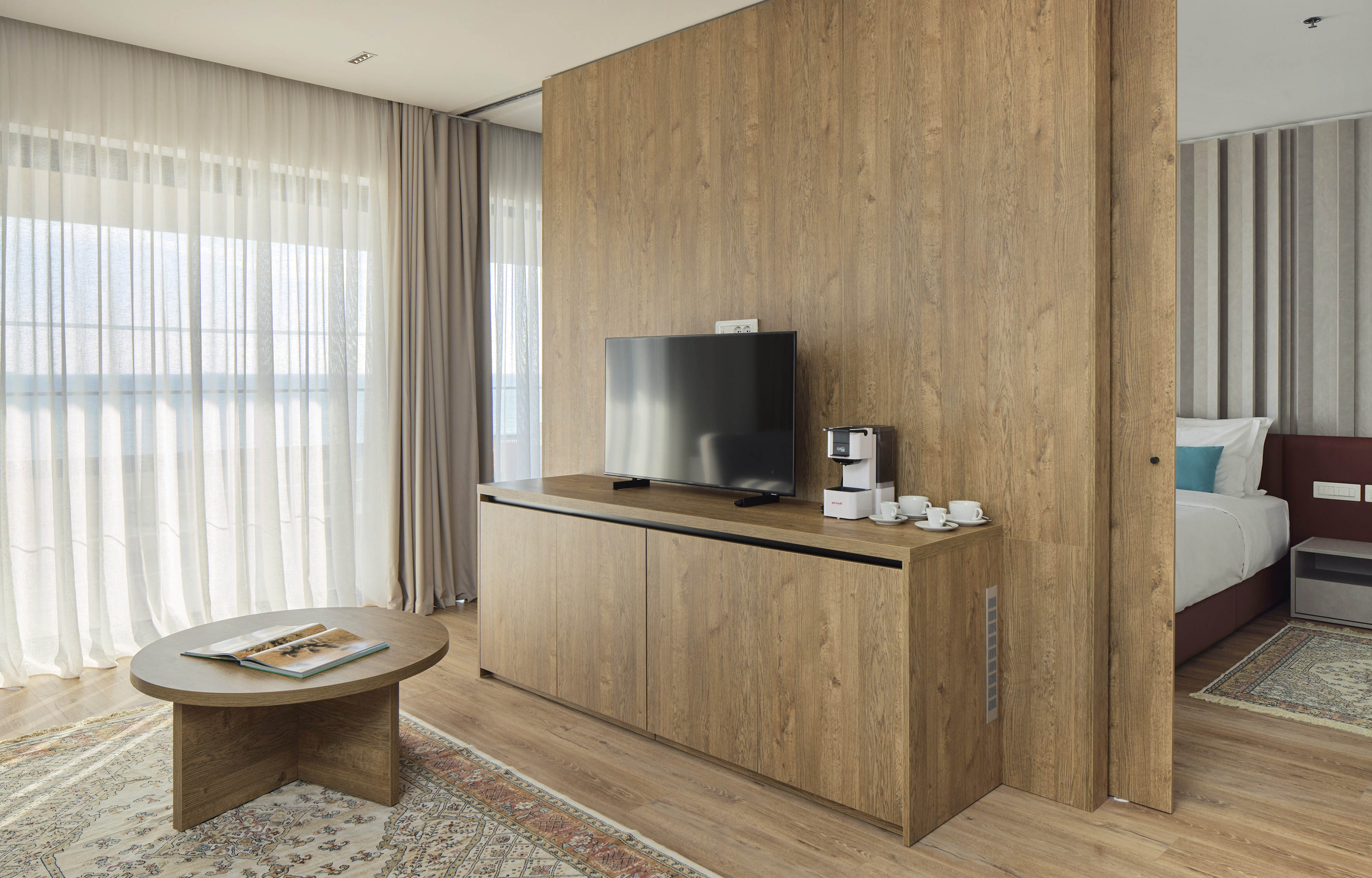 Modern hotel suite with wood paneling, TV, coffee station, and bedroom view.