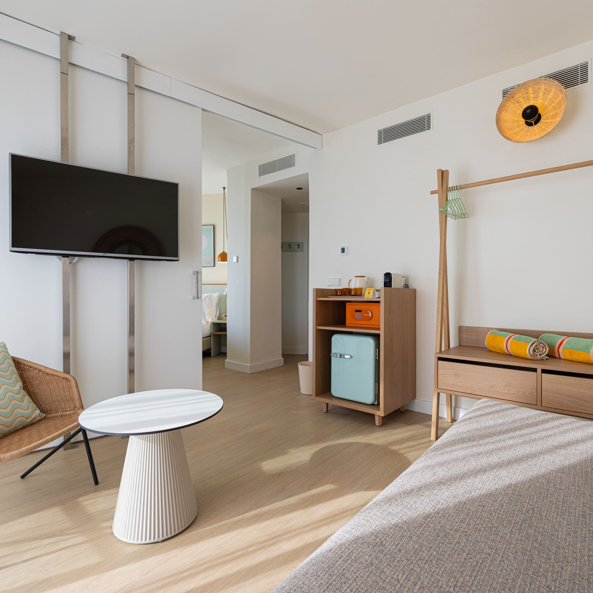 Modern hotel suite with TV, wicker armchair, mini-fridge, wooden rack, colorful towels.