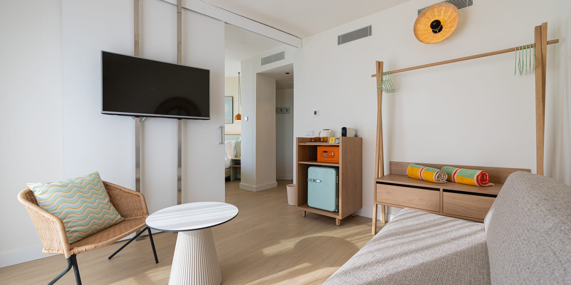 Modern hotel suite with TV, wicker armchair, mini-fridge, wooden rack, colorful towels.