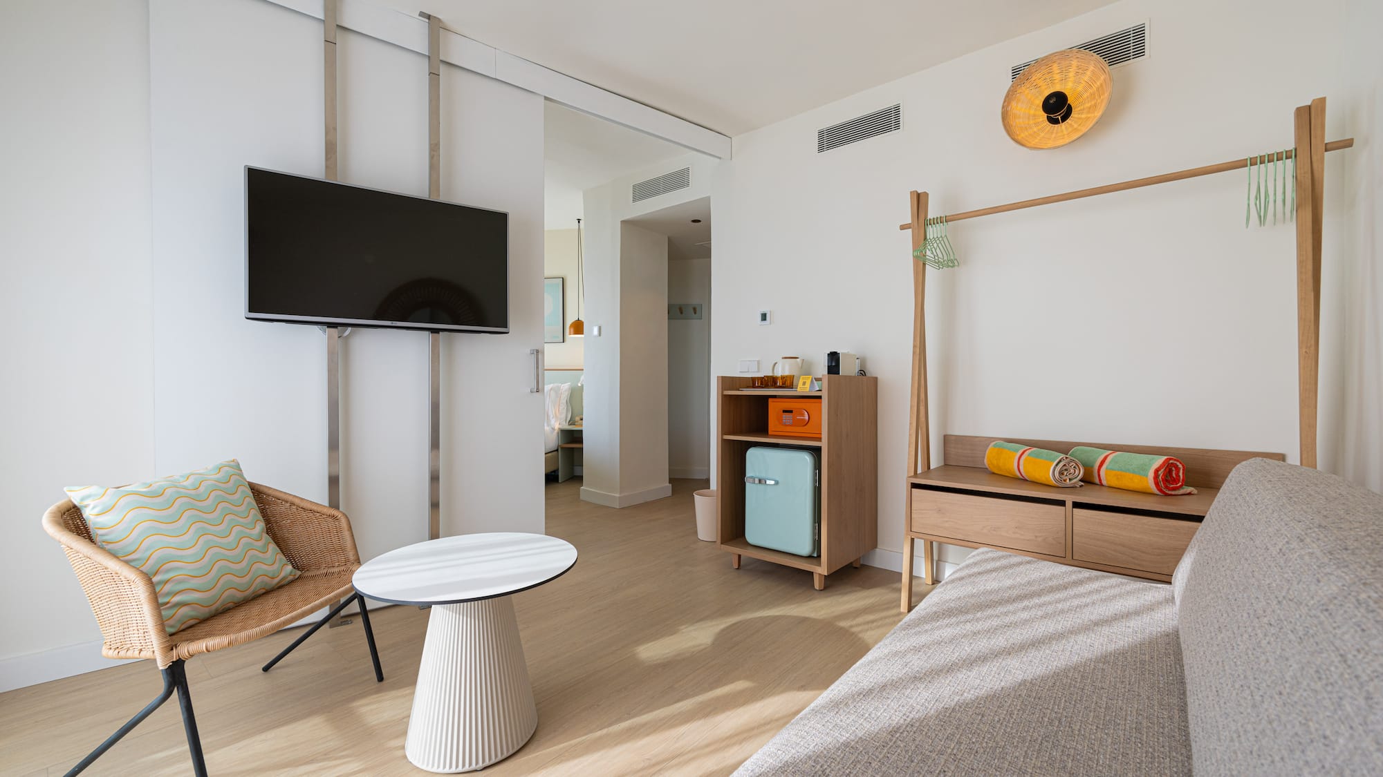 Modern hotel suite with TV, wicker armchair, mini-fridge, wooden rack, colorful towels.