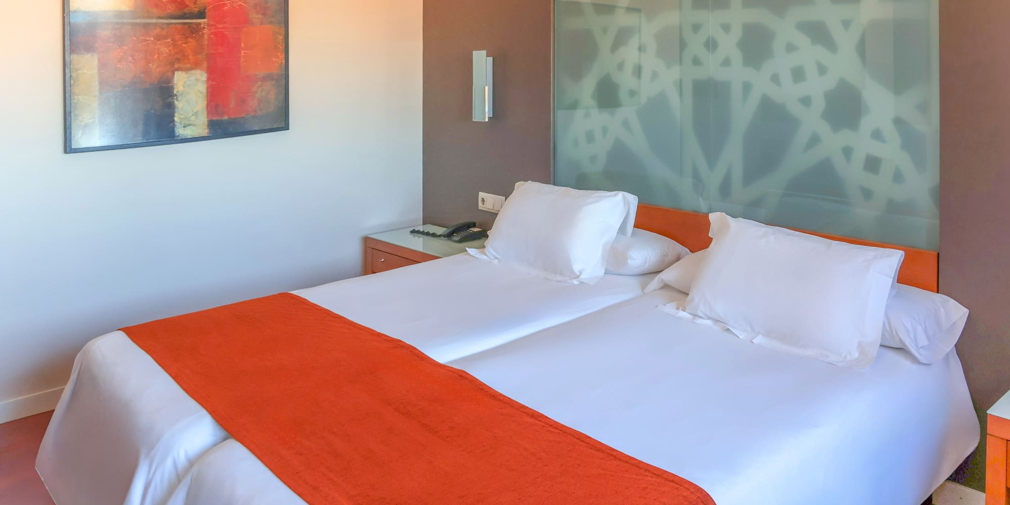 Modern hotel room with twin beds, orange bed runners, and abstract art.