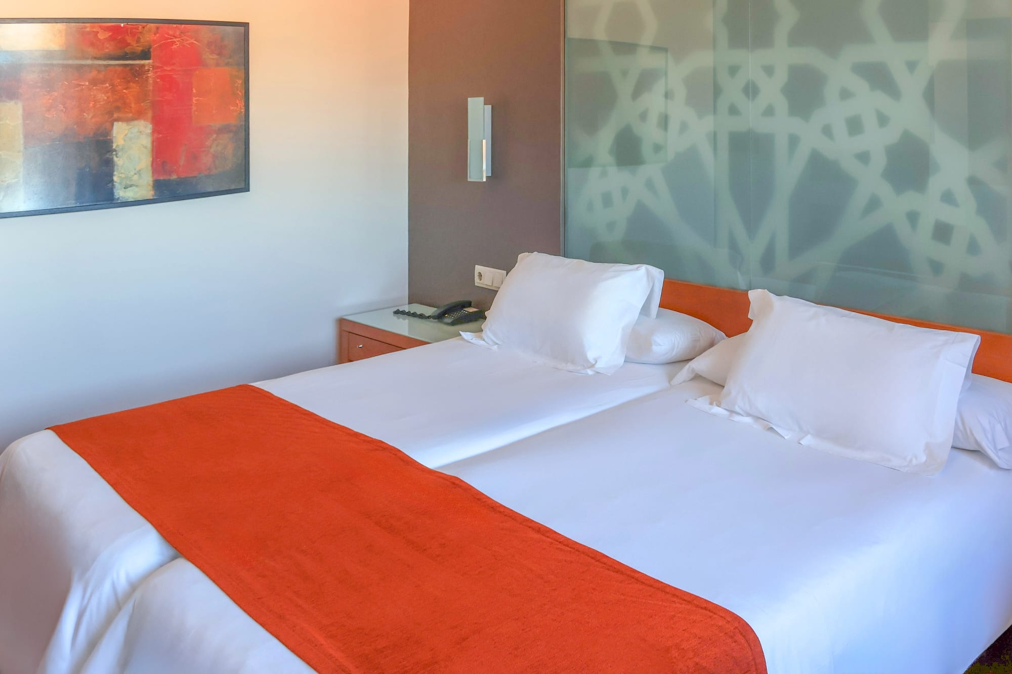 Modern hotel room with twin beds, orange bed runners, and abstract art.