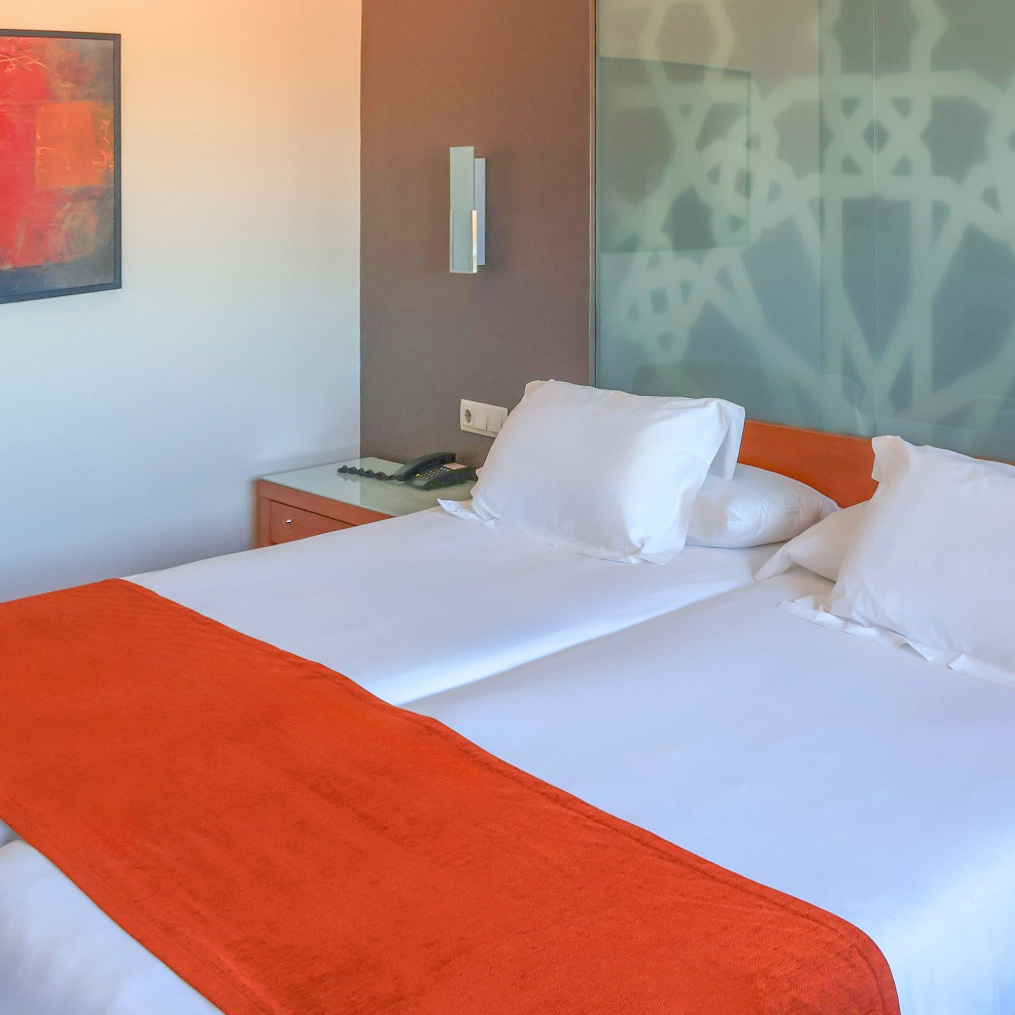 Modern hotel room with twin beds, orange bed runners, and abstract art.