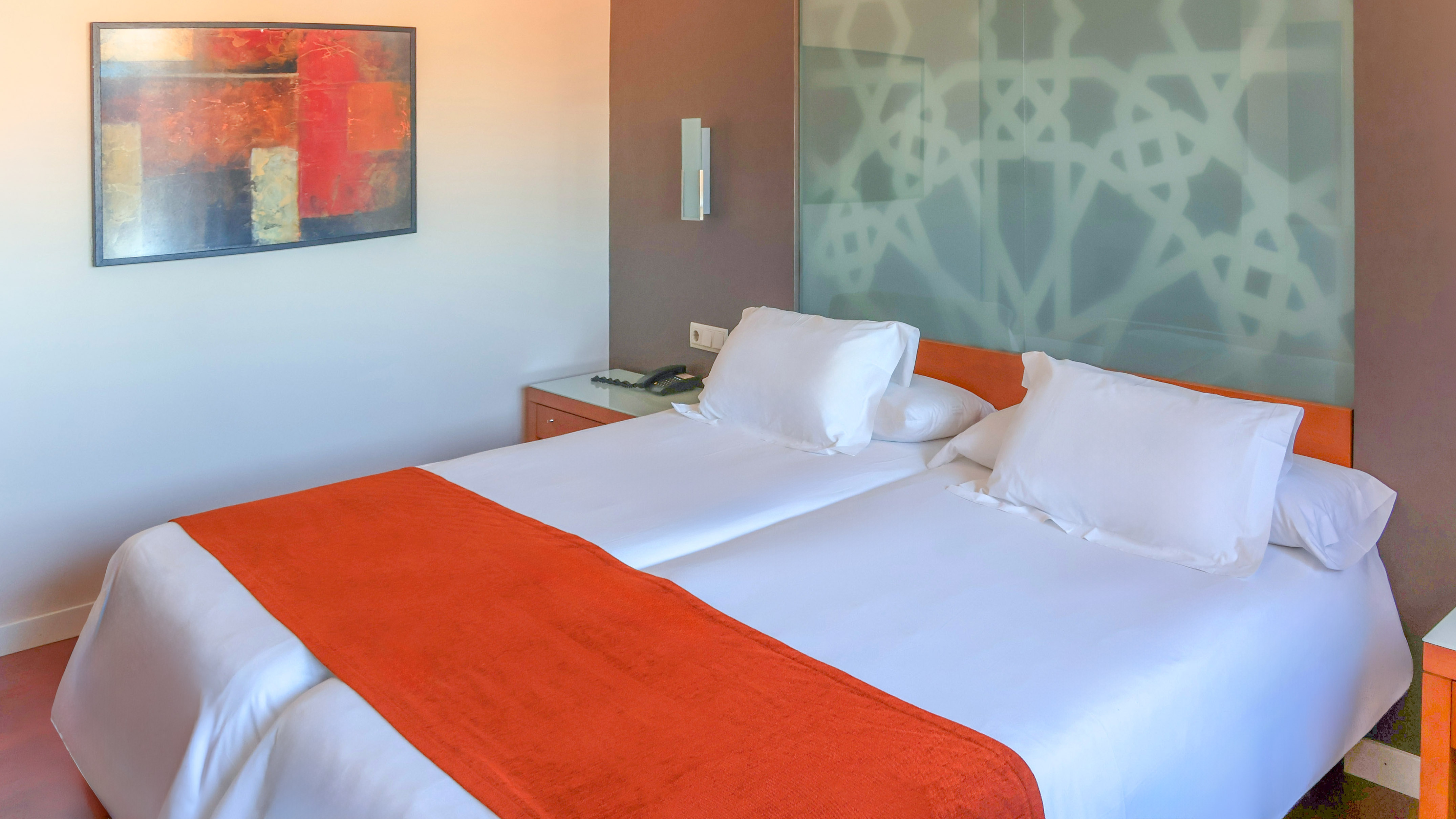 Modern hotel room with twin beds, orange bed runners, and abstract art.