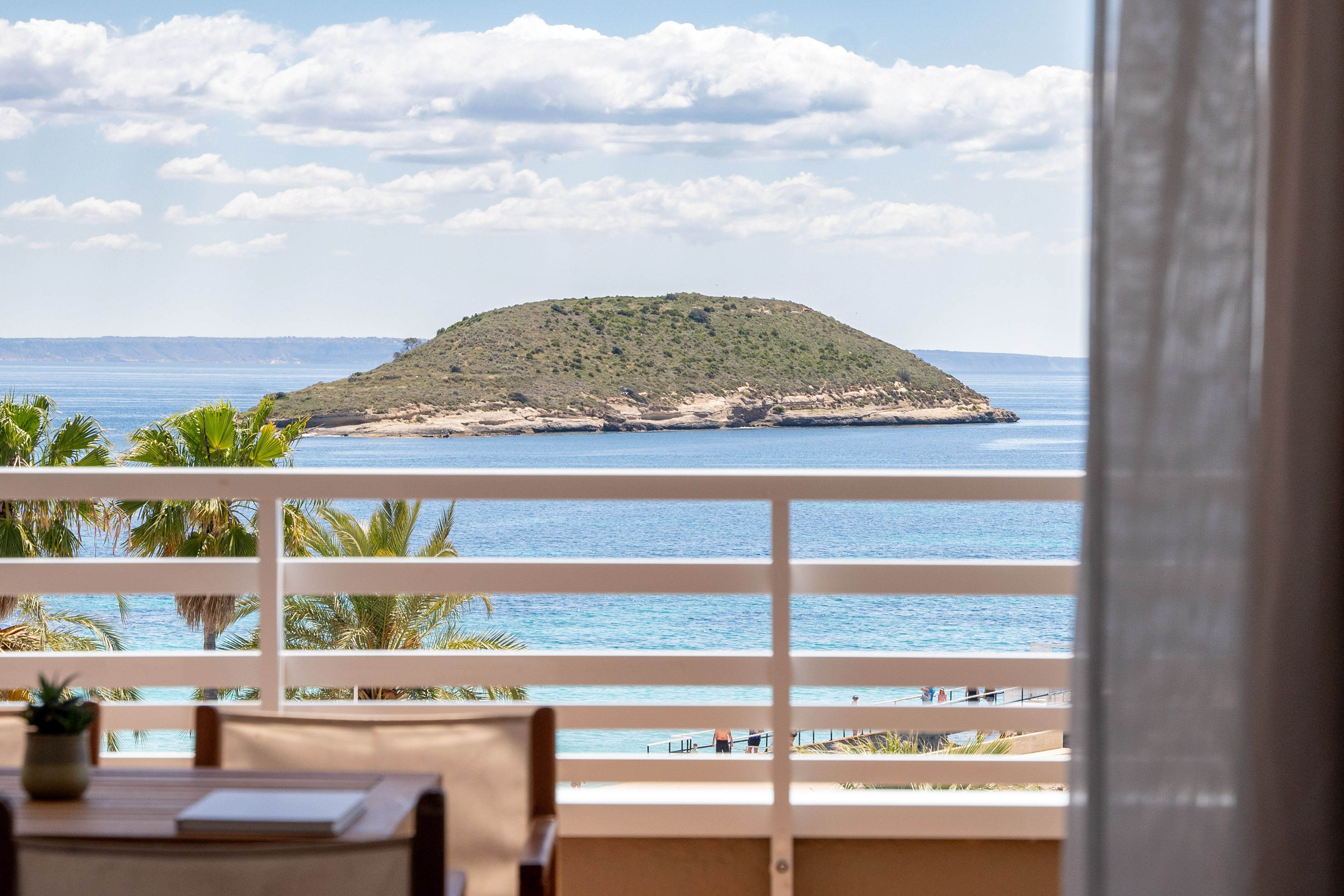 Balcony sea view with green island, blue ocean, palm trees, and white railing.