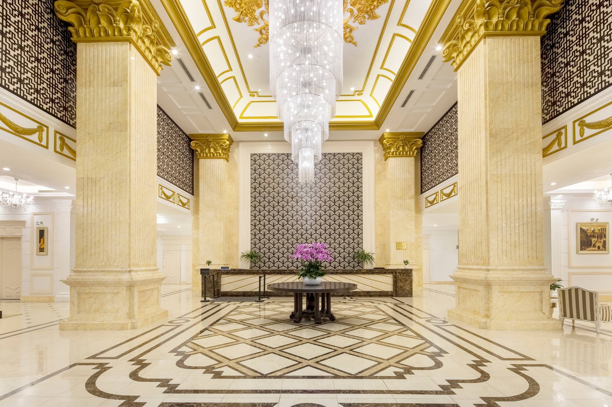 Grand luxury hotel lobby with marble columns, crystal chandelier, and patterned floor.
