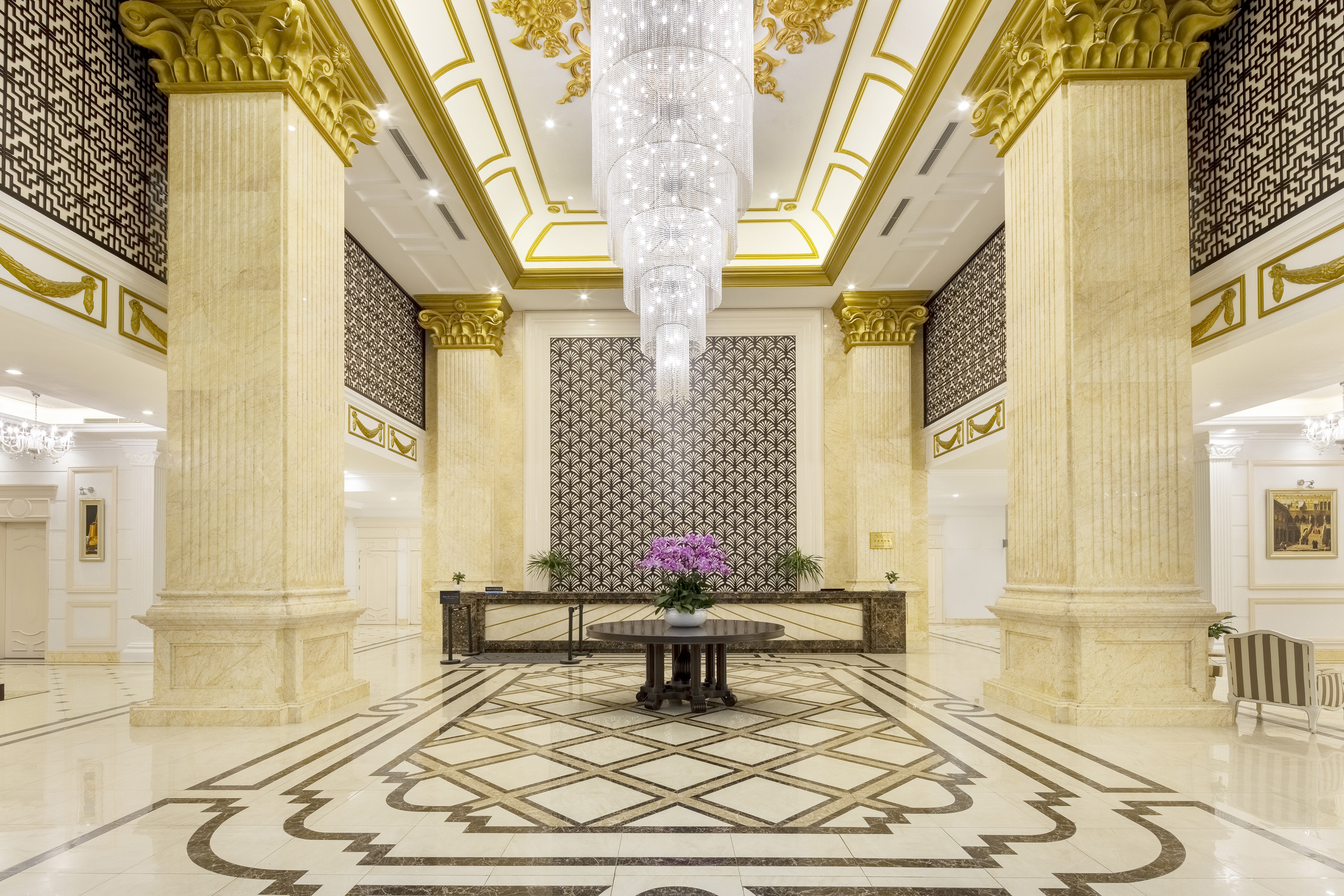 Grand luxury hotel lobby with marble columns, crystal chandelier, and patterned floor.