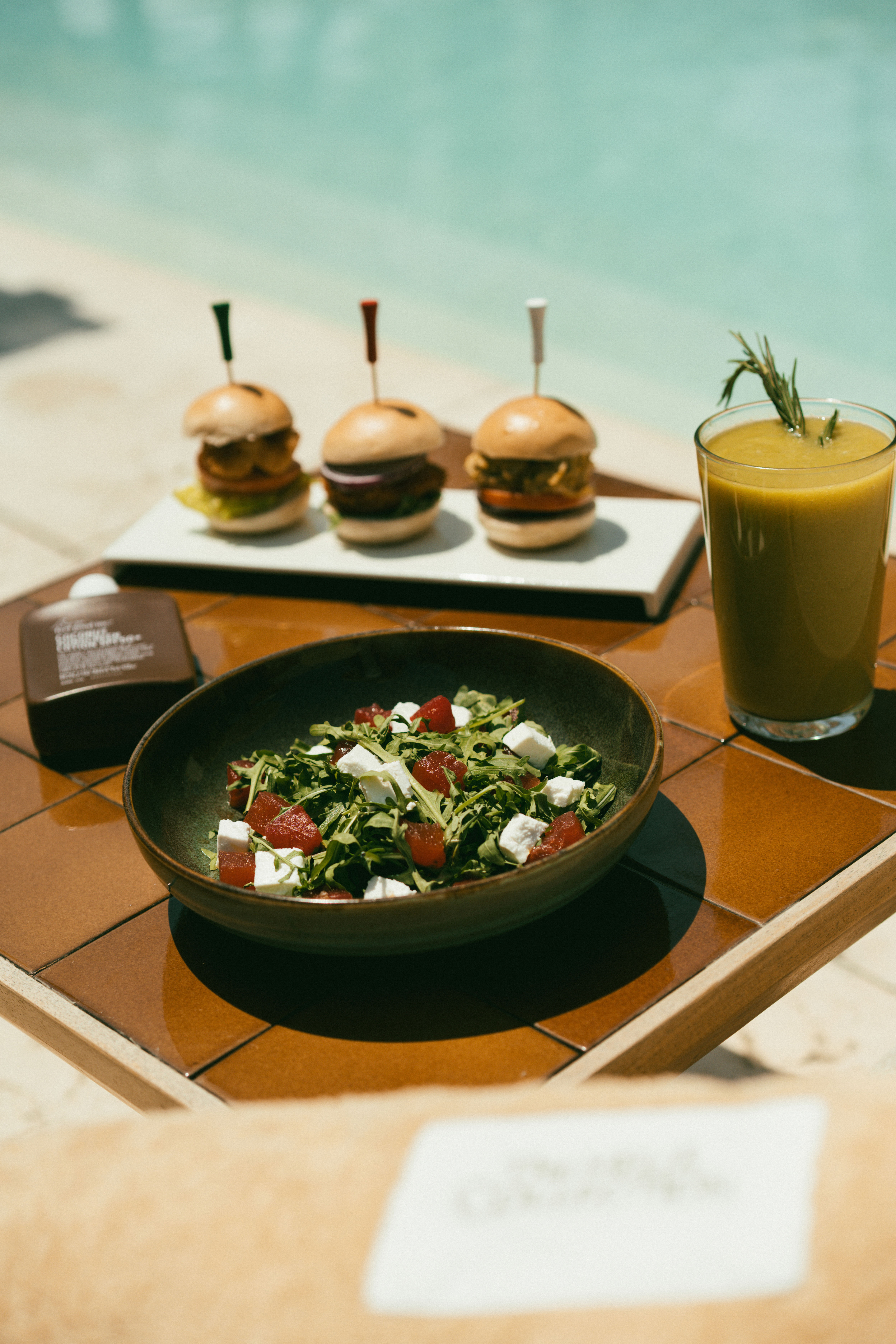 Poolside: mini burgers, arugula feta salad, green juice on tiled table.
