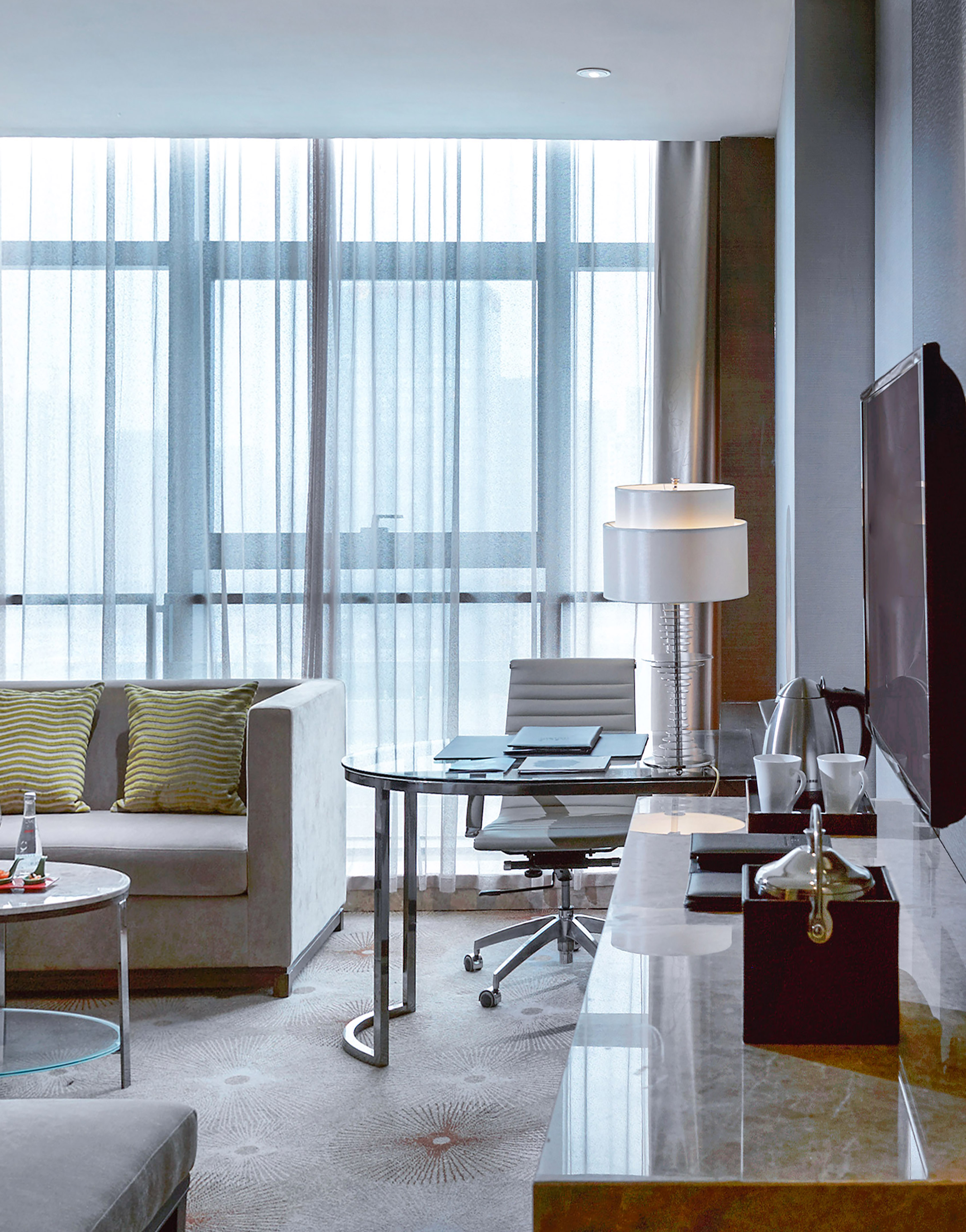 Modern hotel suite with work desk, sofa, and floor-to-ceiling window.