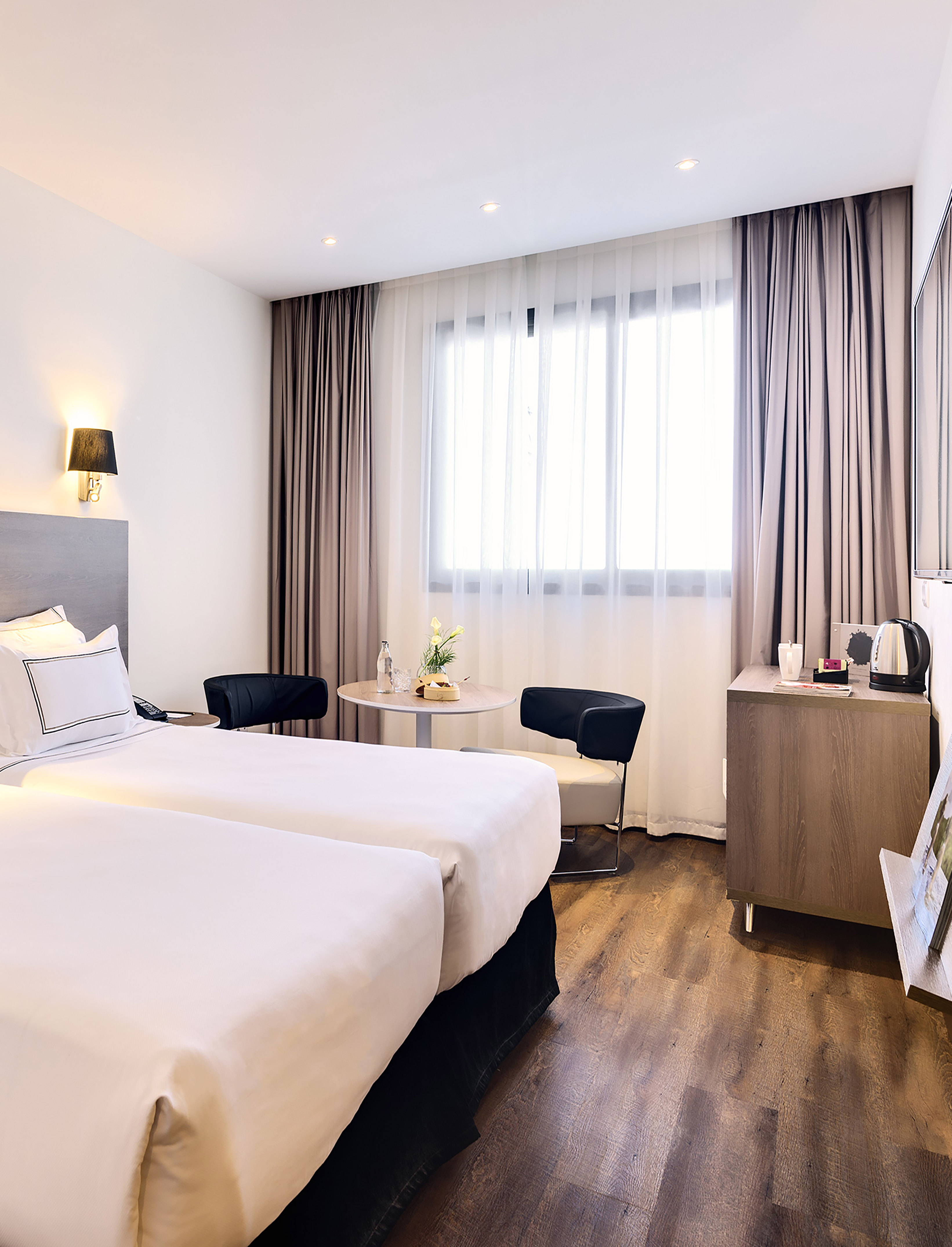 Modern hotel room with twin beds, wooden floor, and bright window.