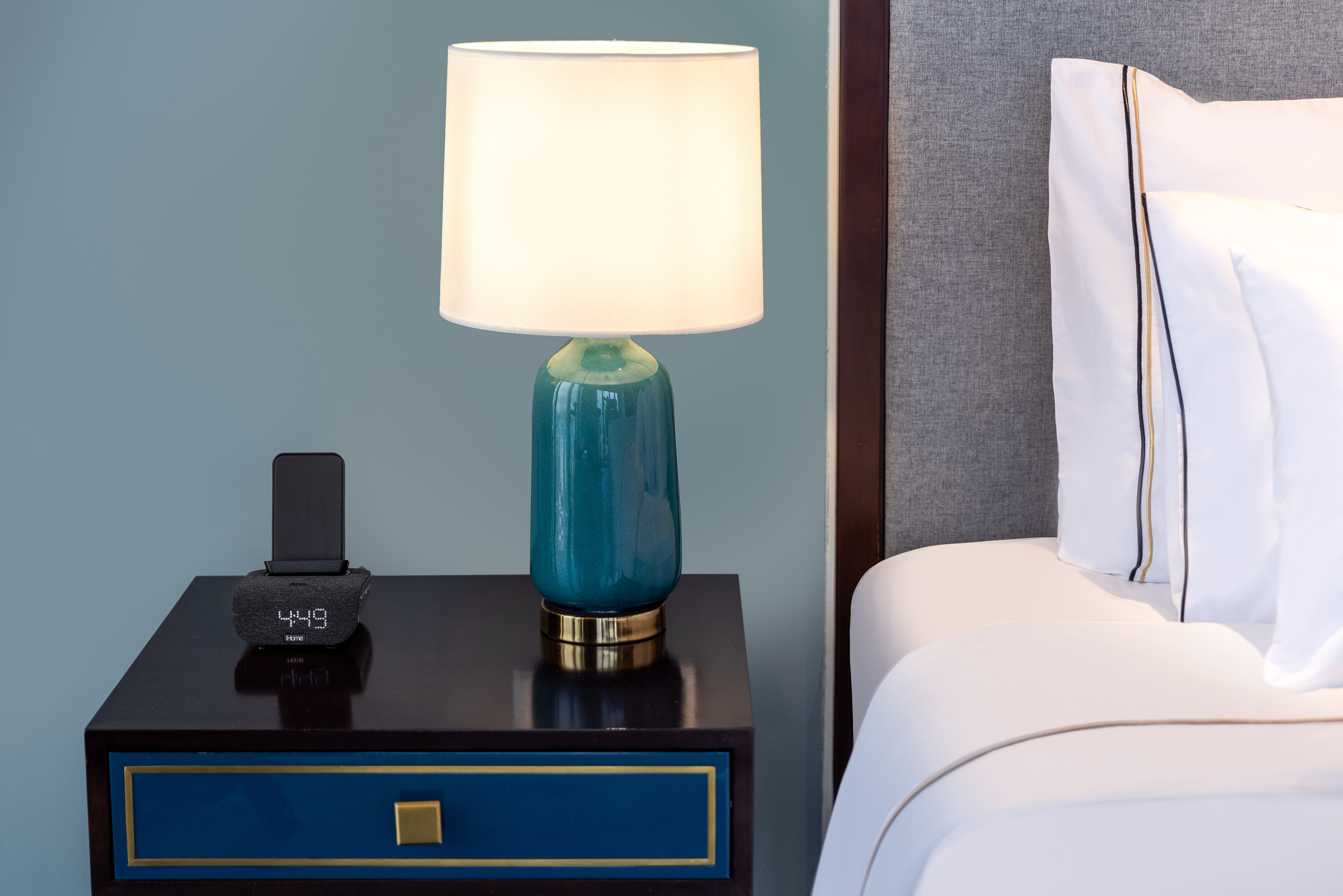 Hotel bedroom nightstand with teal lamp, iHome clock, and white bed.