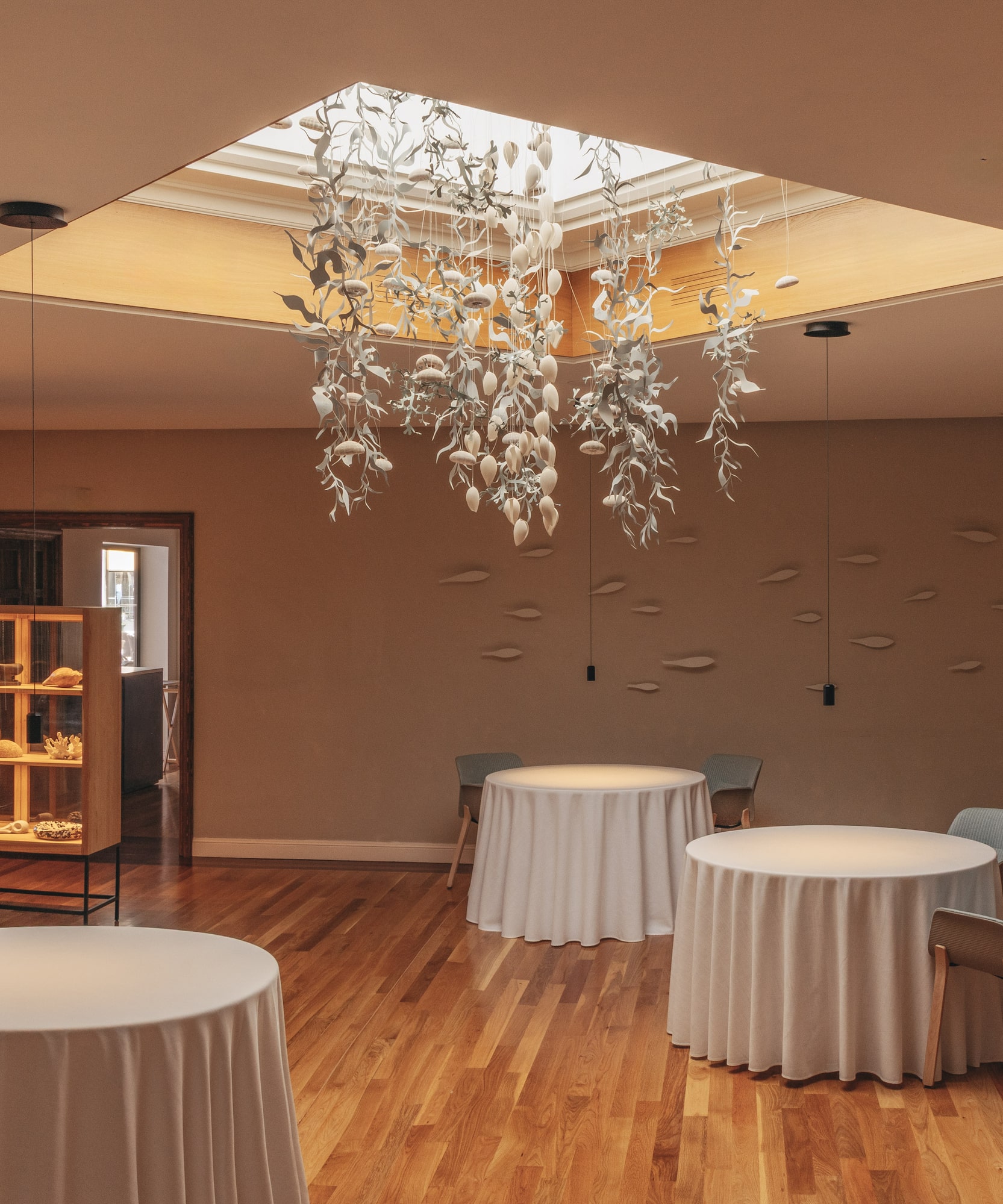 Elegant restaurant interior: leaf chandelier, skylight, white tables, wooden floor.