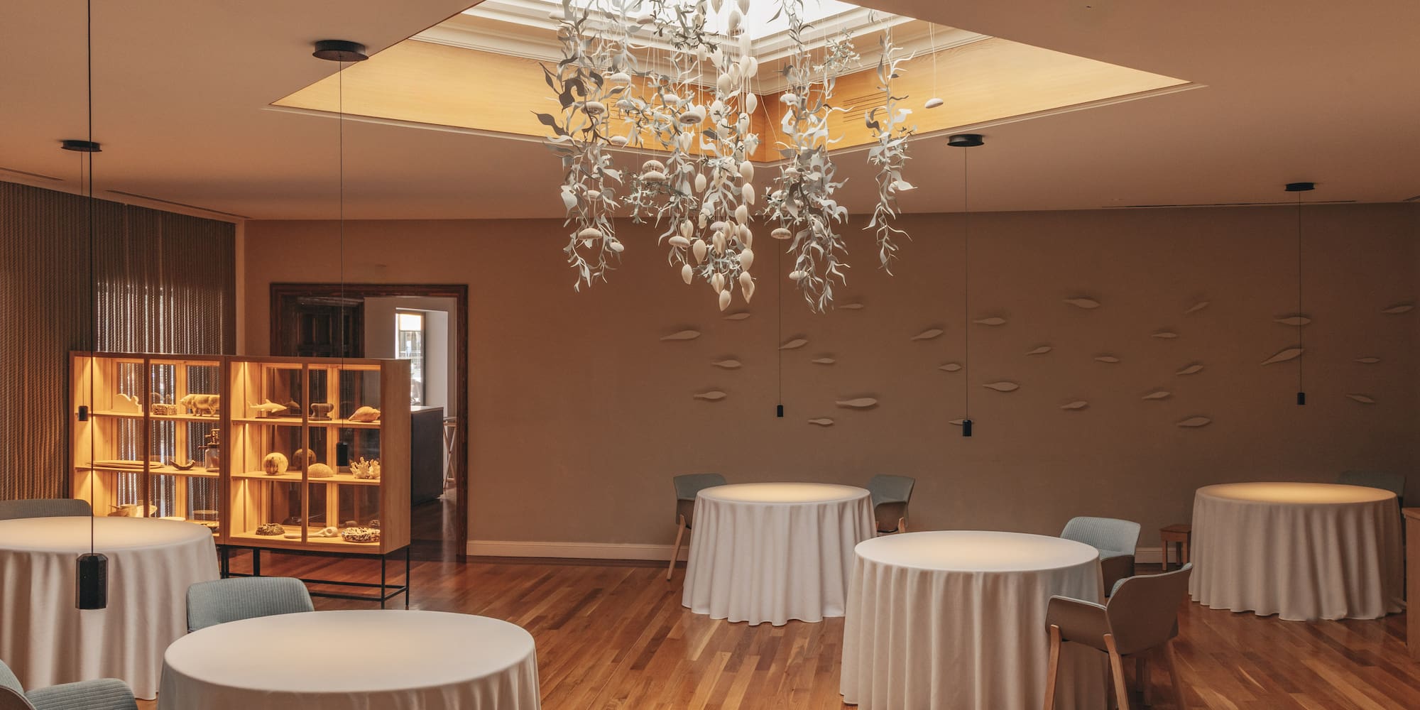 Elegant restaurant interior: leaf chandelier, skylight, white tables, wooden floor.