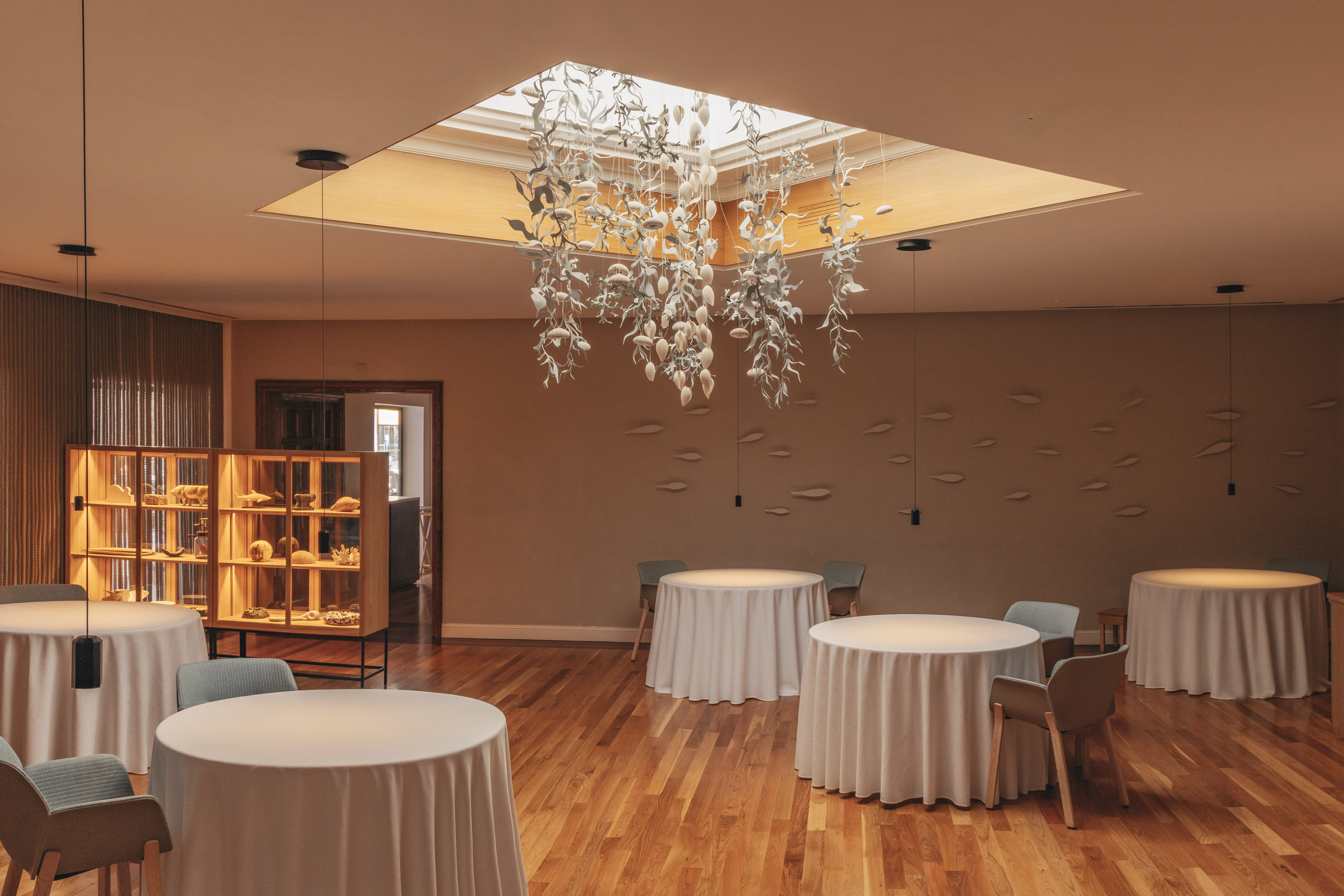Elegant restaurant interior: leaf chandelier, skylight, white tables, wooden floor.