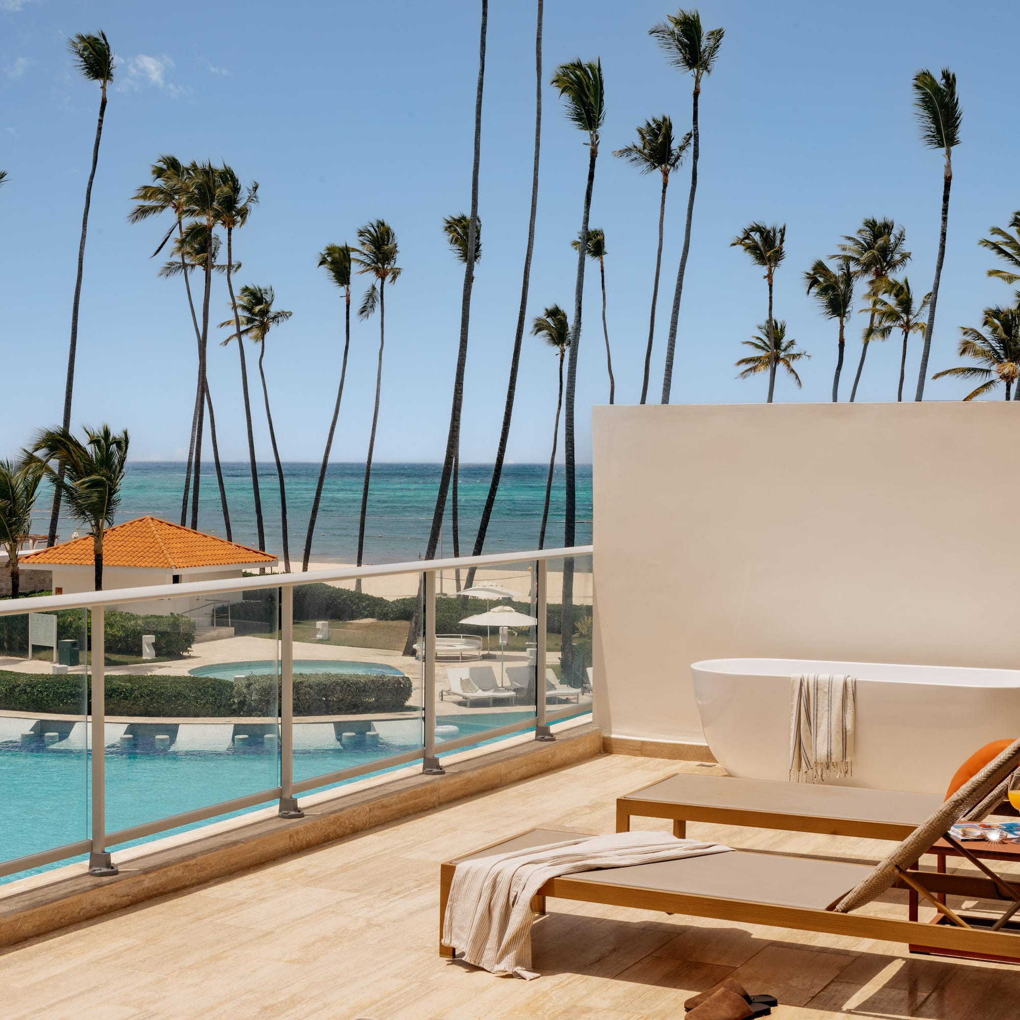 Oceanfront resort balcony with outdoor tub, lounge chairs, pool, and palm trees.
