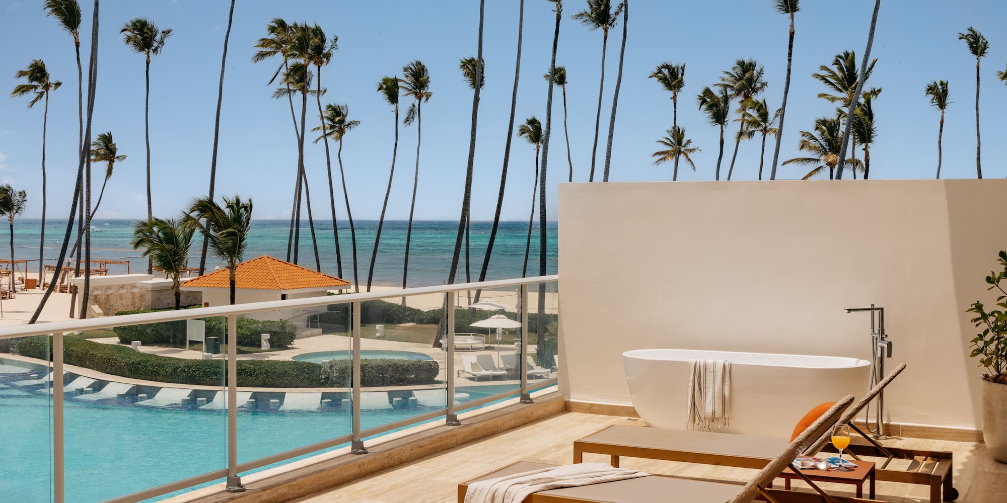Oceanfront resort balcony with outdoor tub, lounge chairs, pool, and palm trees.
