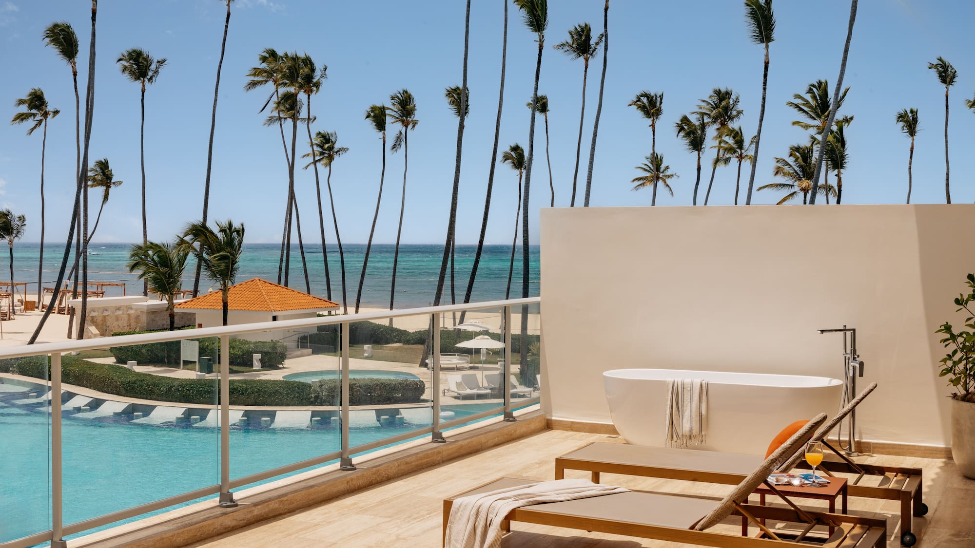 Oceanfront resort balcony with outdoor tub, lounge chairs, pool, and palm trees.