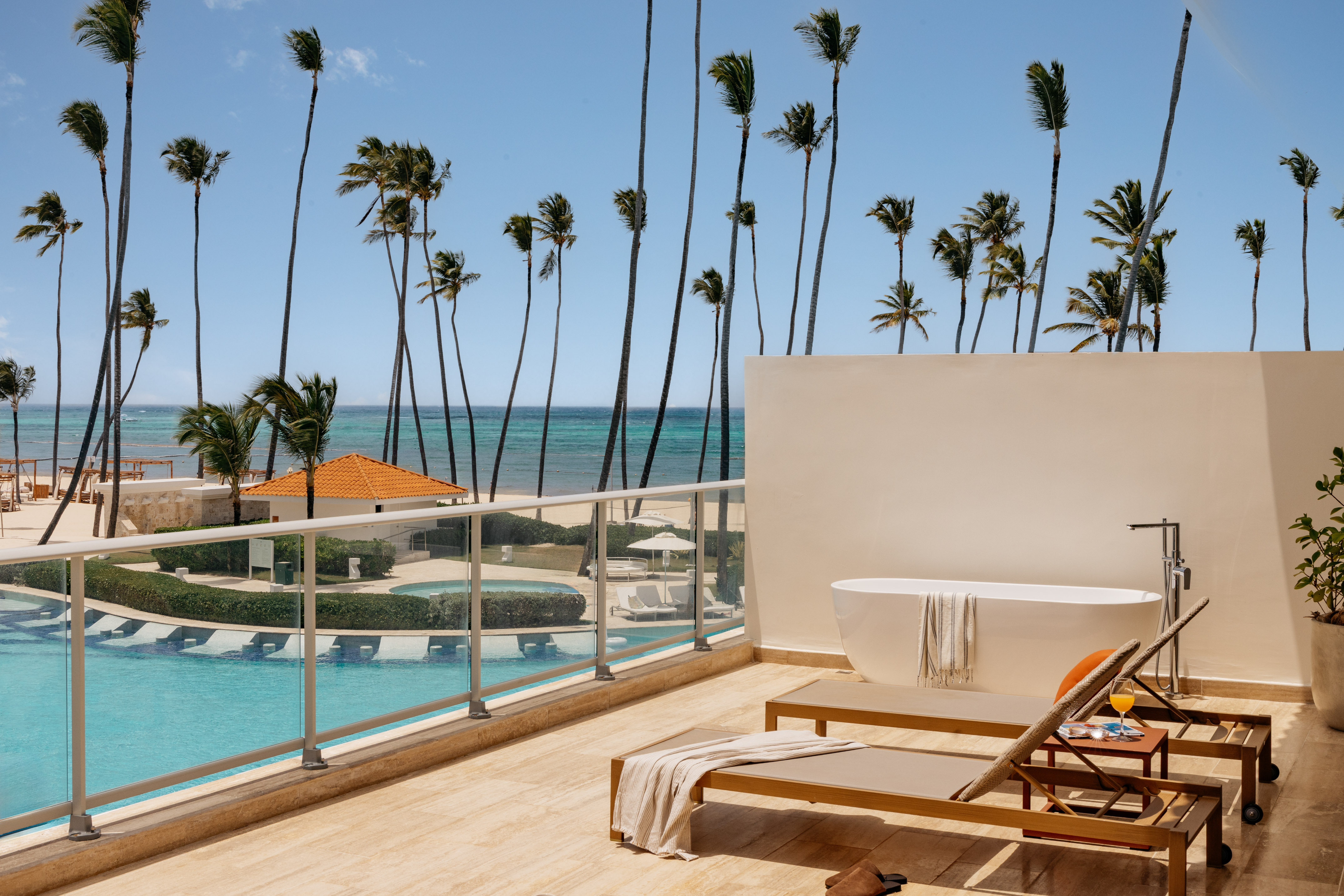 Oceanfront resort balcony with outdoor tub, lounge chairs, pool, and palm trees.