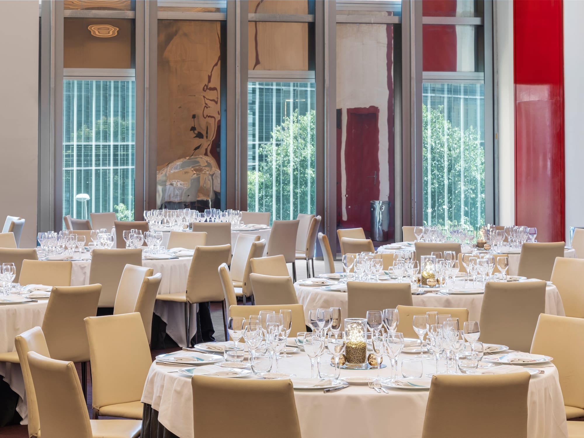 Salón de banquetes con mesas redondas, sillas claras y amplios ventanales.