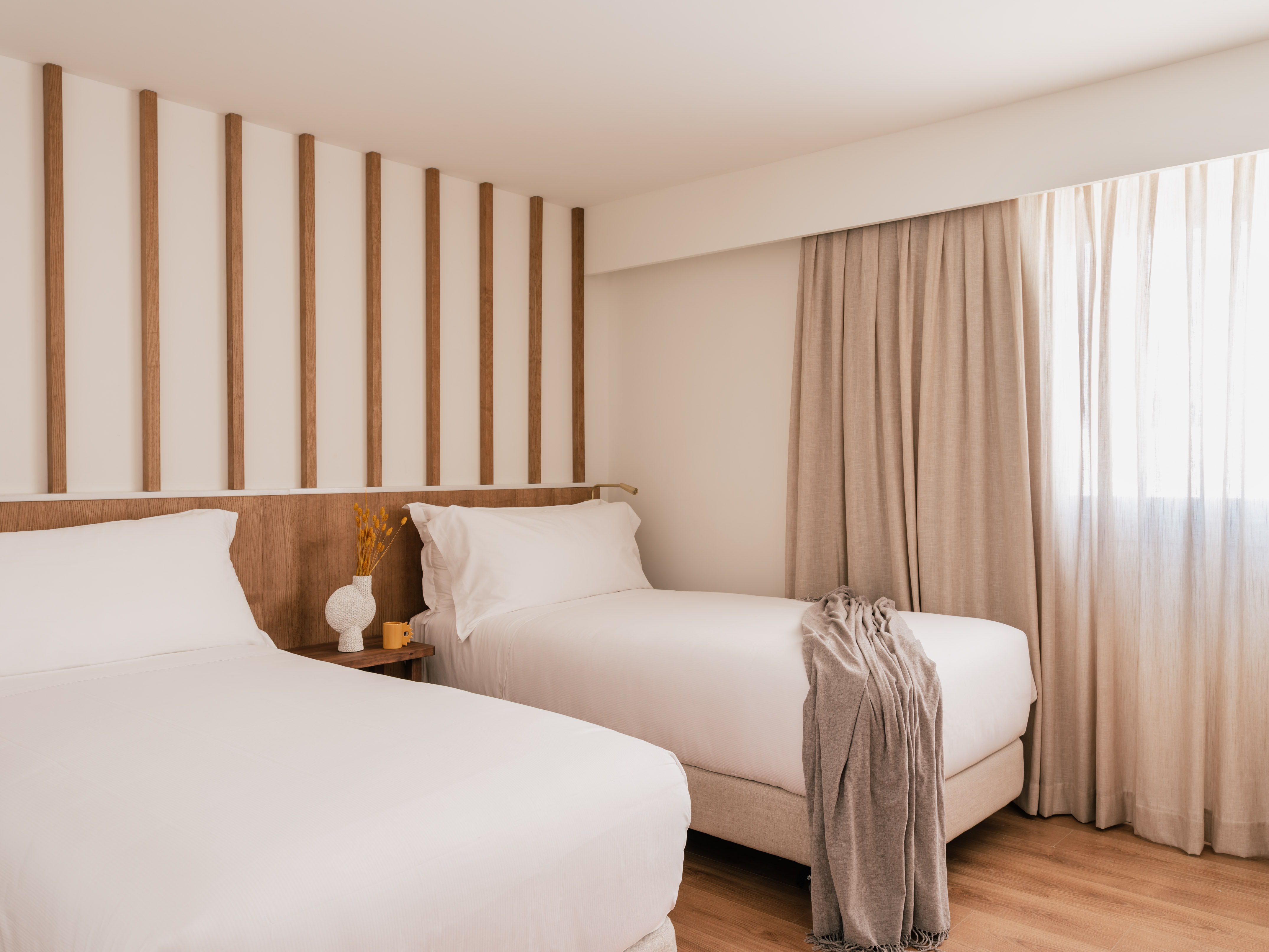 Modern hotel room with twin beds, white bedding, wood paneling, and light curtains.