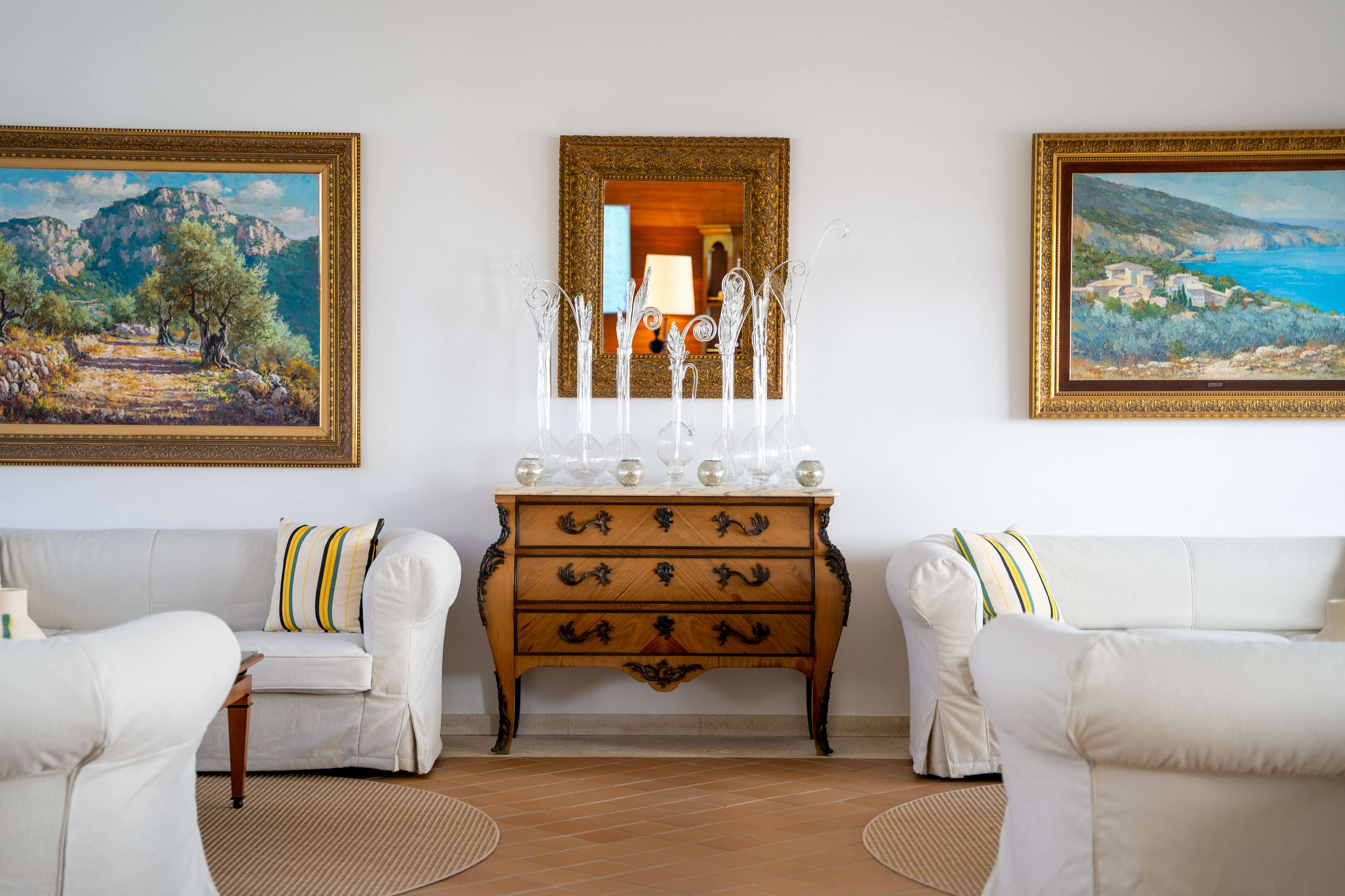 Elegant room: white sofas, antique dresser, mirror, and two landscape paintings.