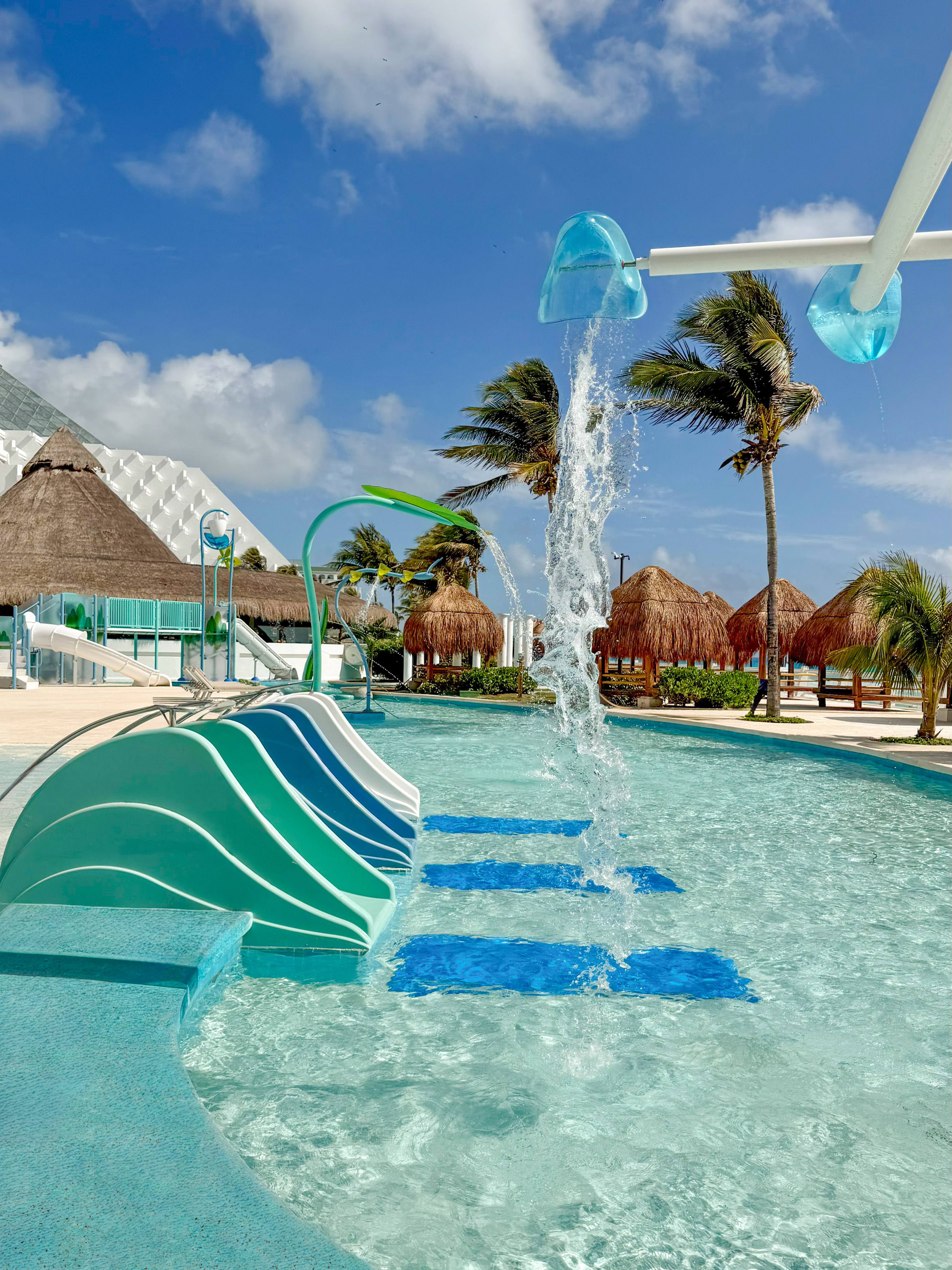 Resort water park with blue slides, splash features, palm trees, and huts.