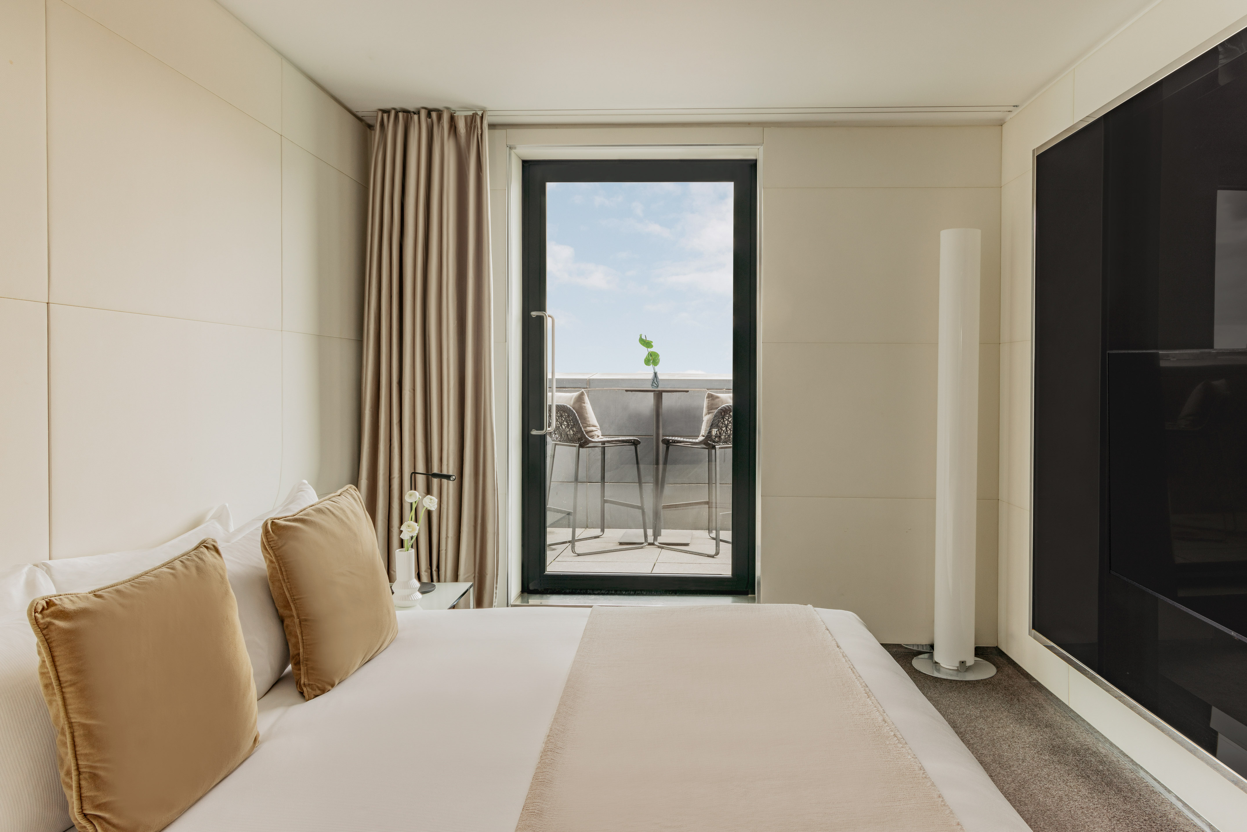 Modern hotel bedroom with large bed, balcony seating, and minimalist decor.