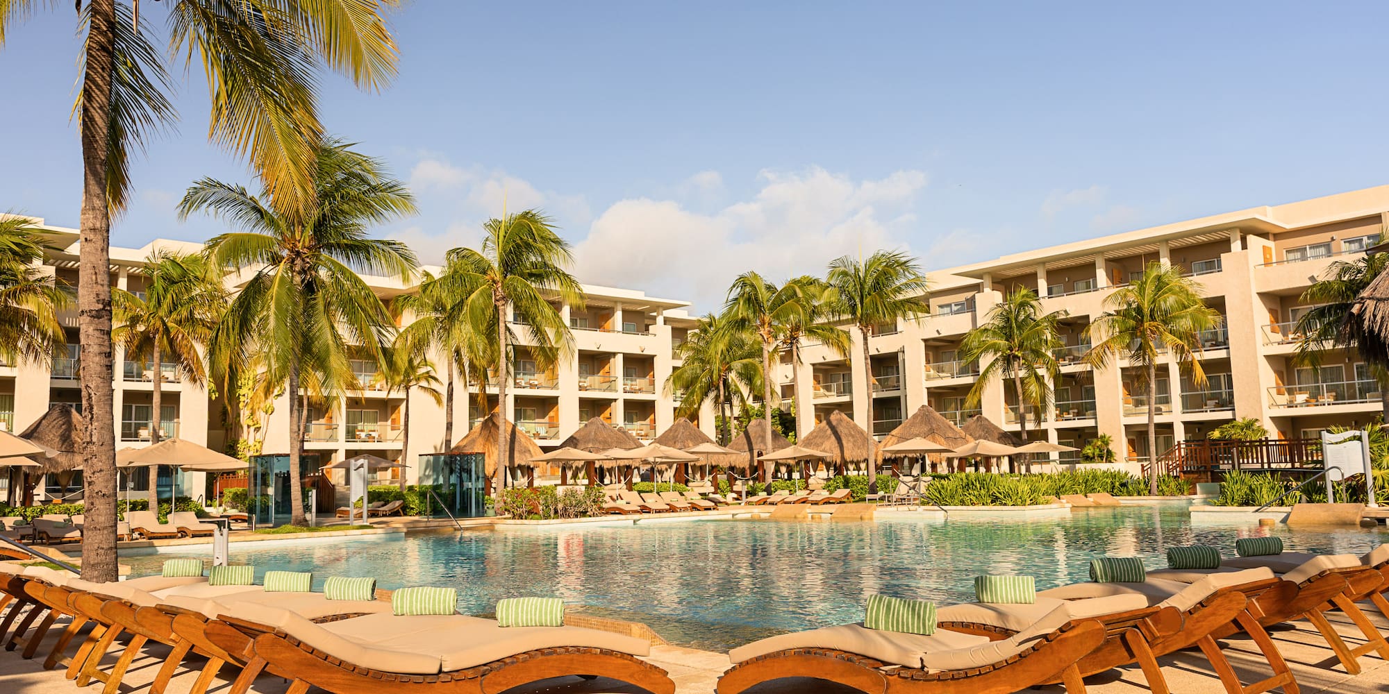 Tropical resort pool, lounge chairs, palm trees, and hotel buildings.