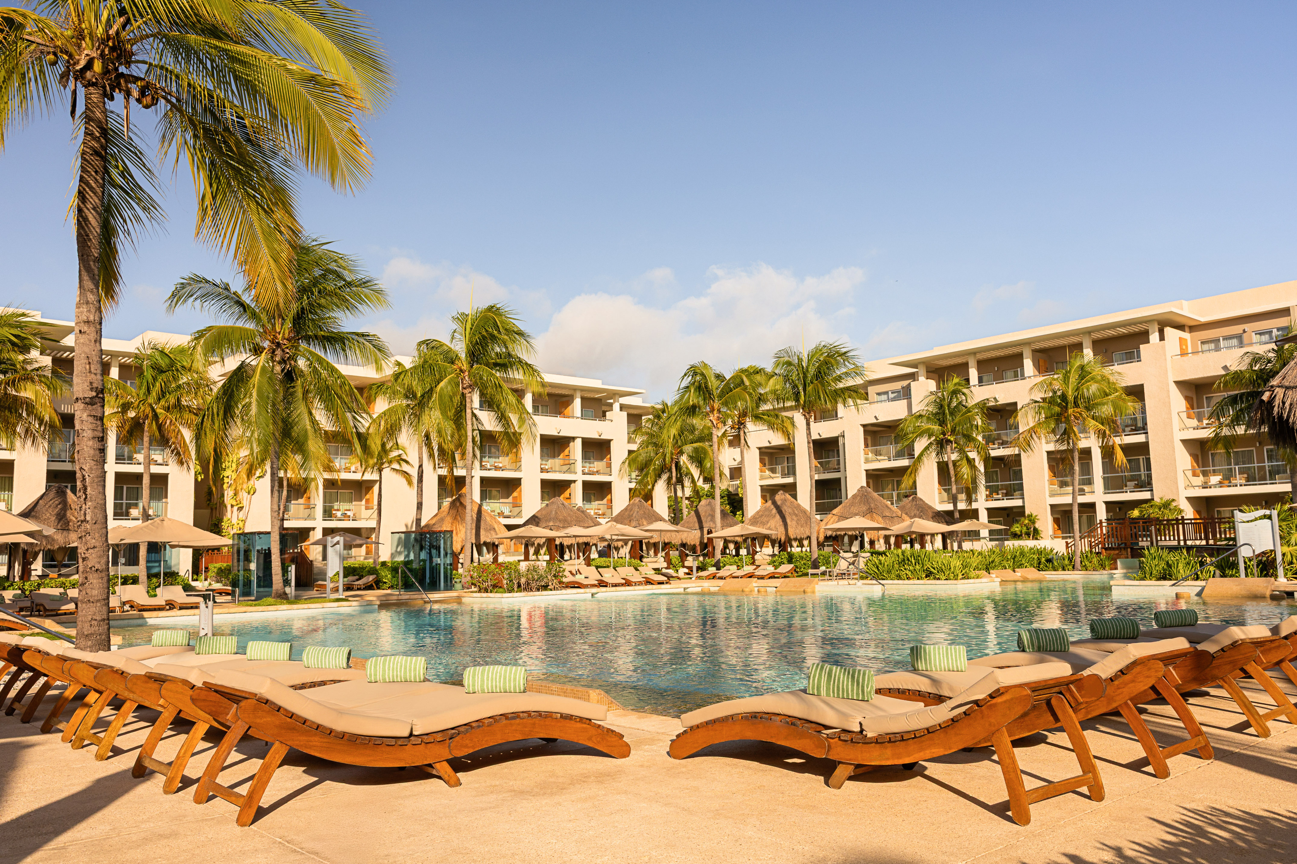 Tropical resort pool, lounge chairs, palm trees, and hotel buildings.