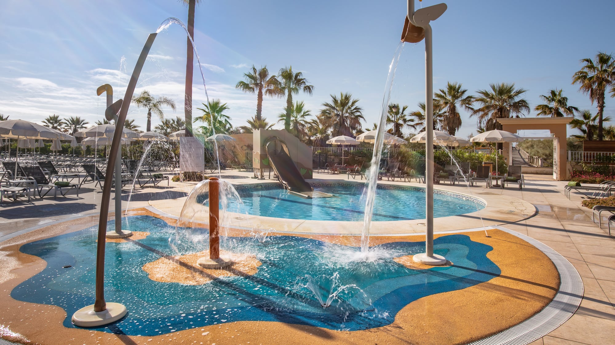 Sunny resort splash pad with water fountains, slide, pool, and palm trees.