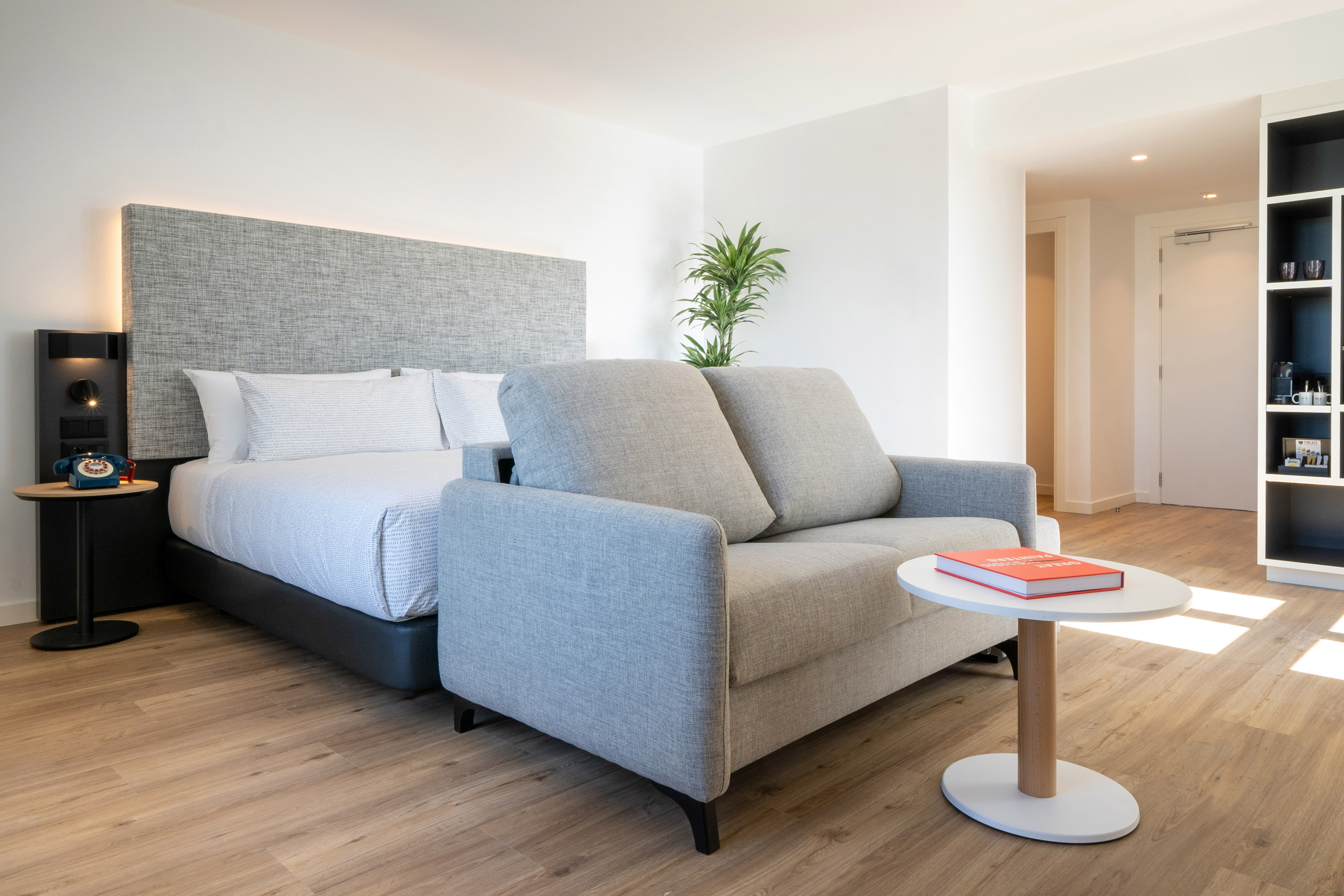 Modern hotel room with bed, grey sofa, wooden floor, and minimalist decor.