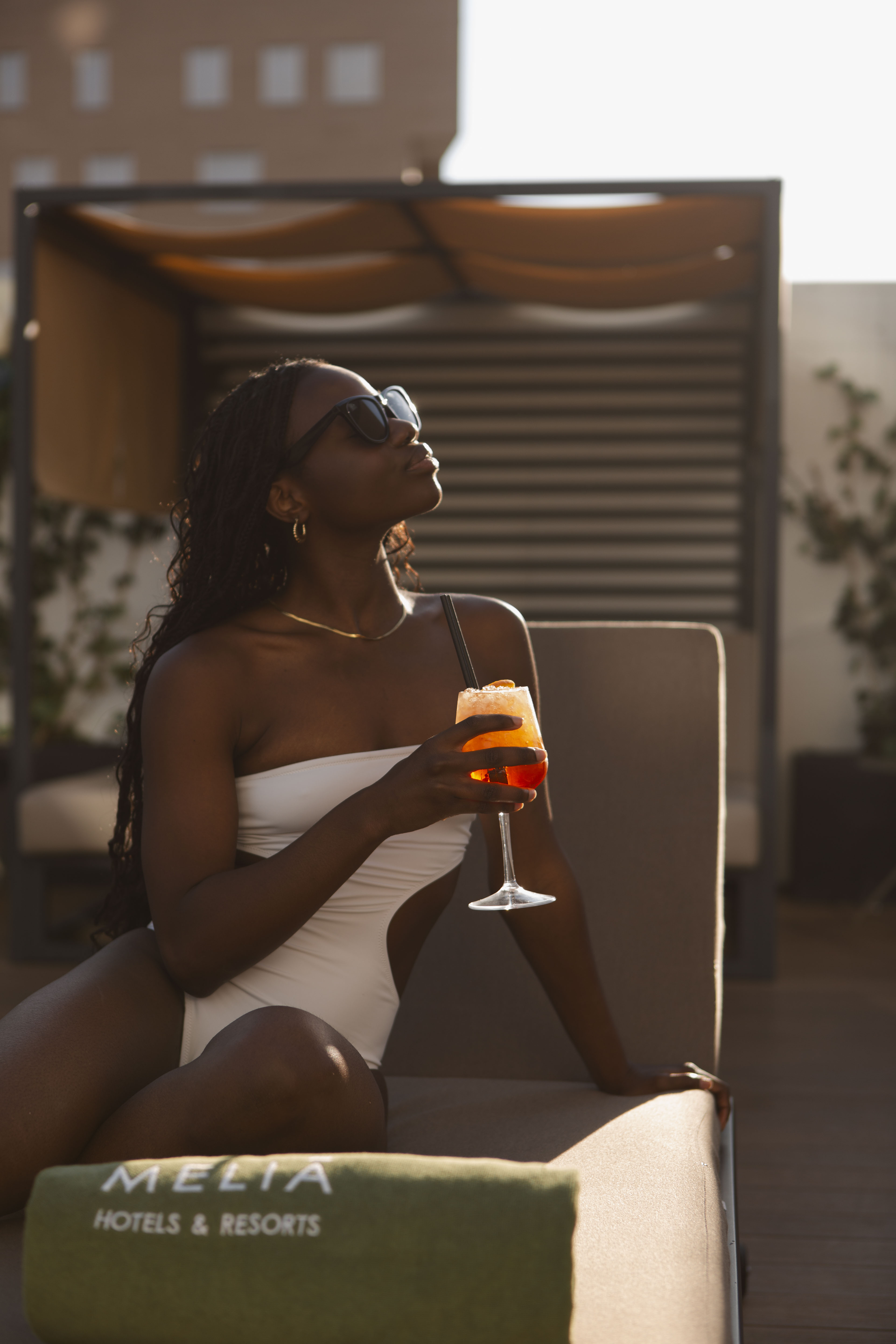 Black woman in white swimsuit, sunglasses, holding cocktail at Melia resort.