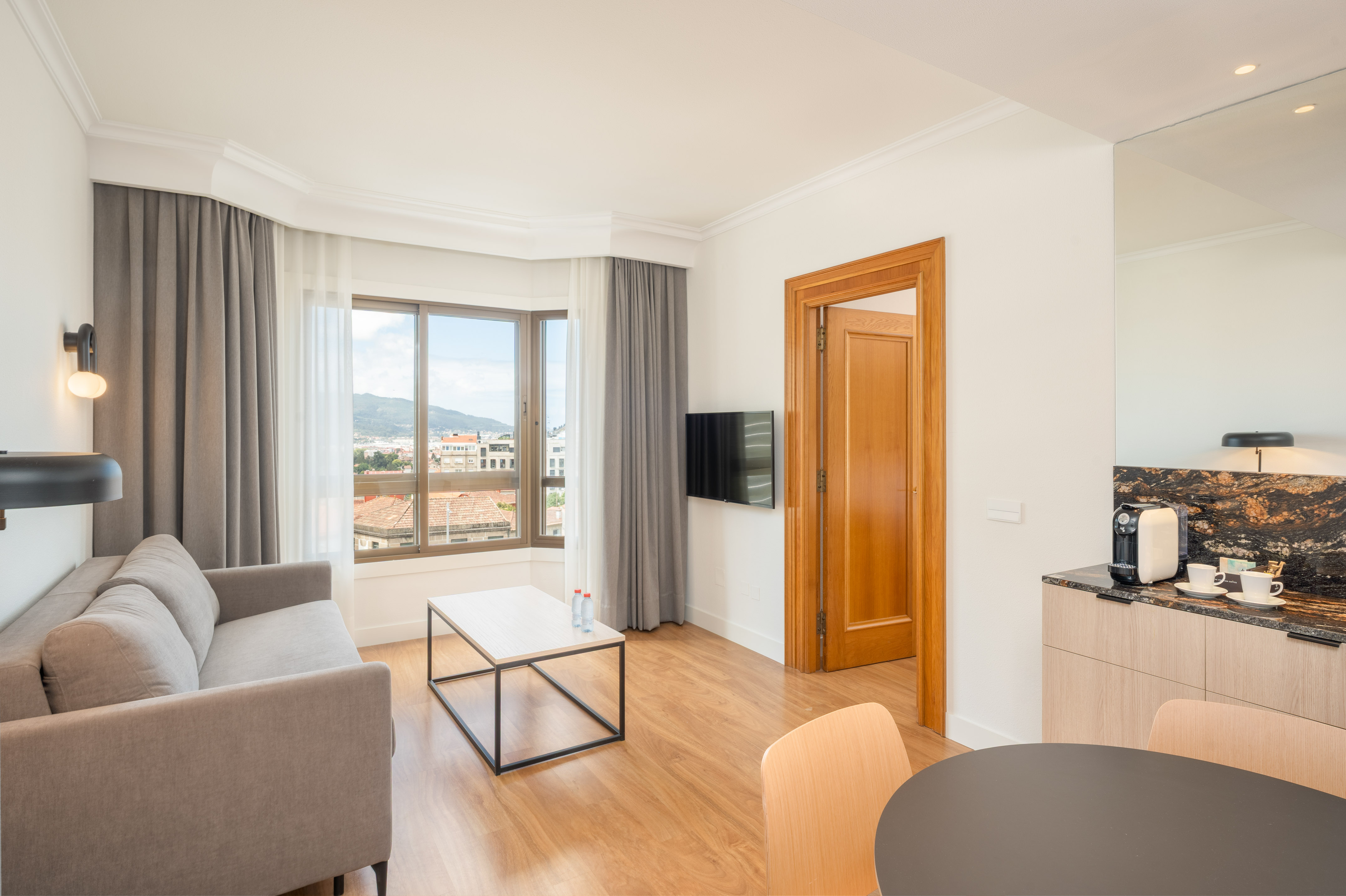 Contemporary hotel suite living area with sofa, city view, and kitchenette.