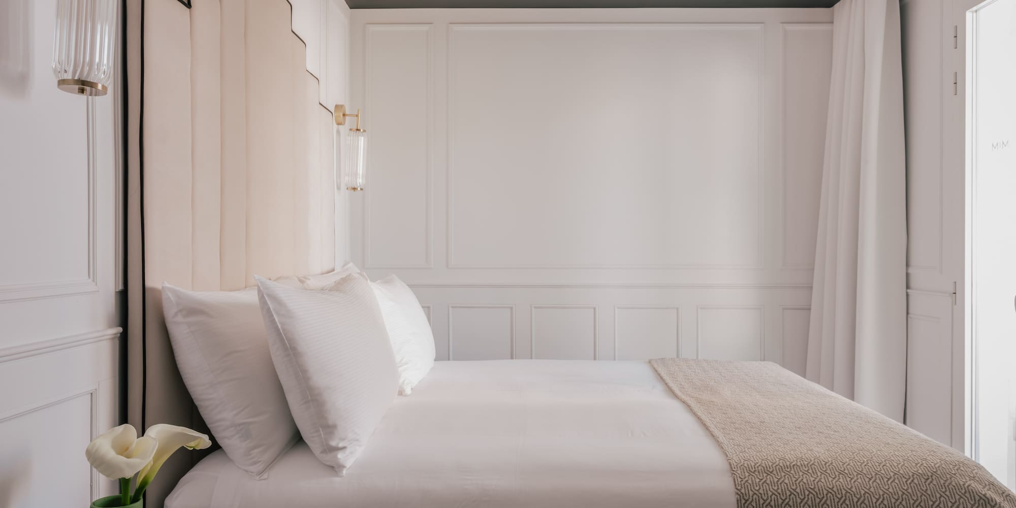Luxury hotel bedroom with white bed, beige headboard, and gold wall sconces.