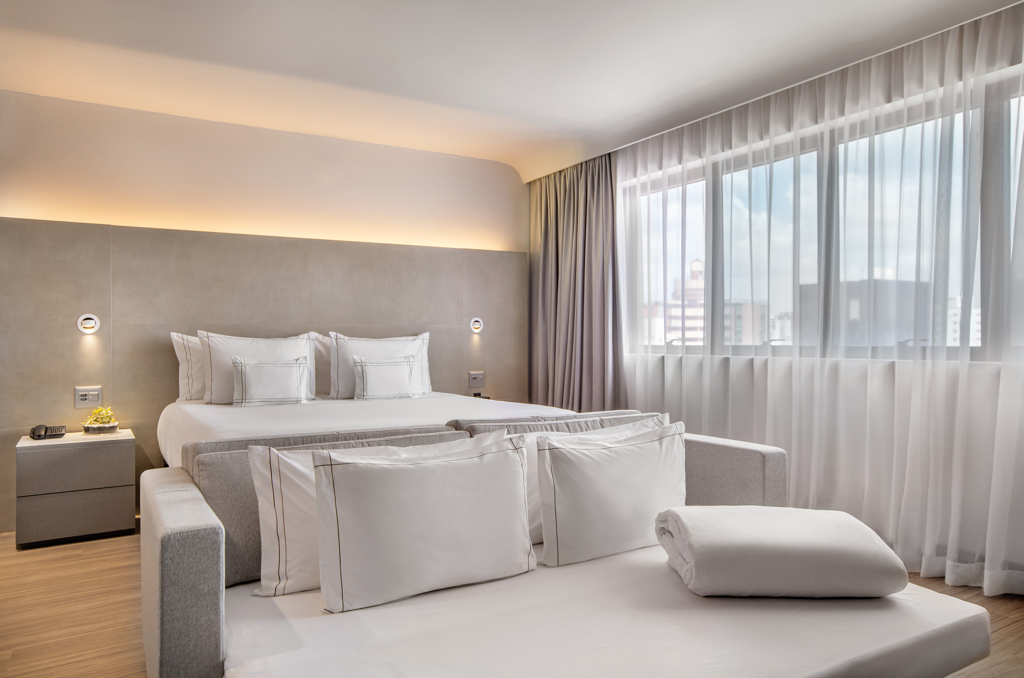Modern hotel room with king bed, sofa, neutral decor, and sheer windows.
