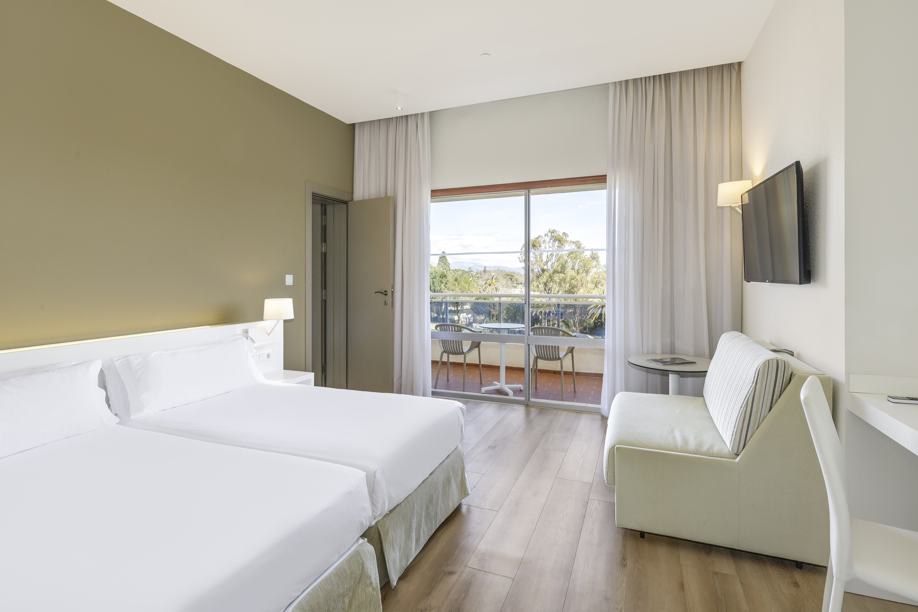Bright hotel room with twin beds, balcony, flat-screen TV, and scenic outdoor view.