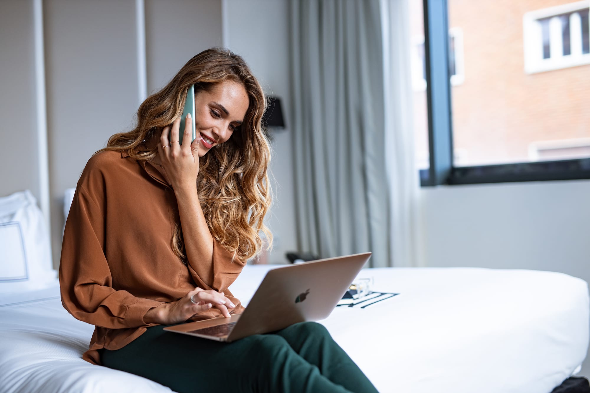 a woman sitting on a bed with a laptop and phone