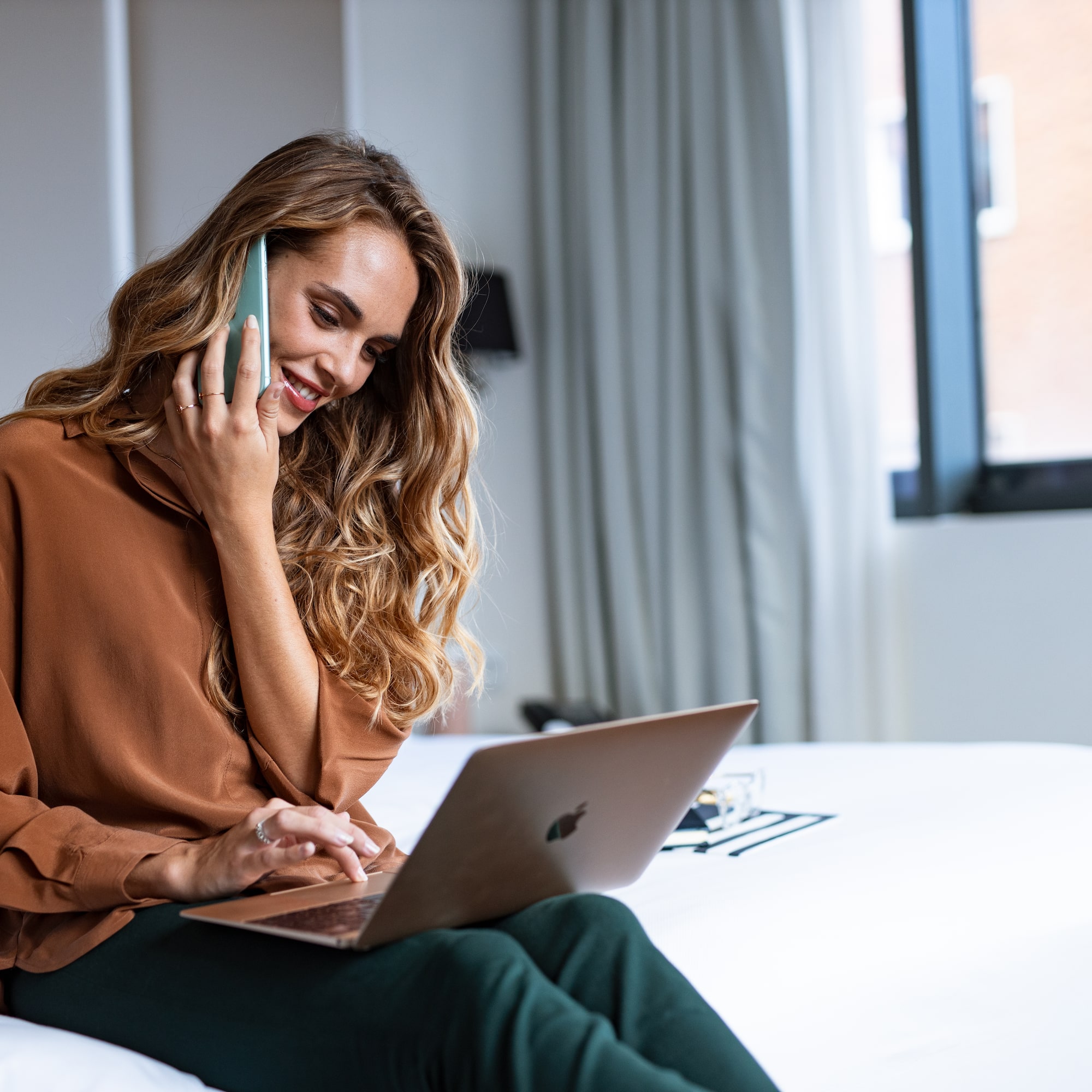 a woman sitting on a bed with a laptop and phone