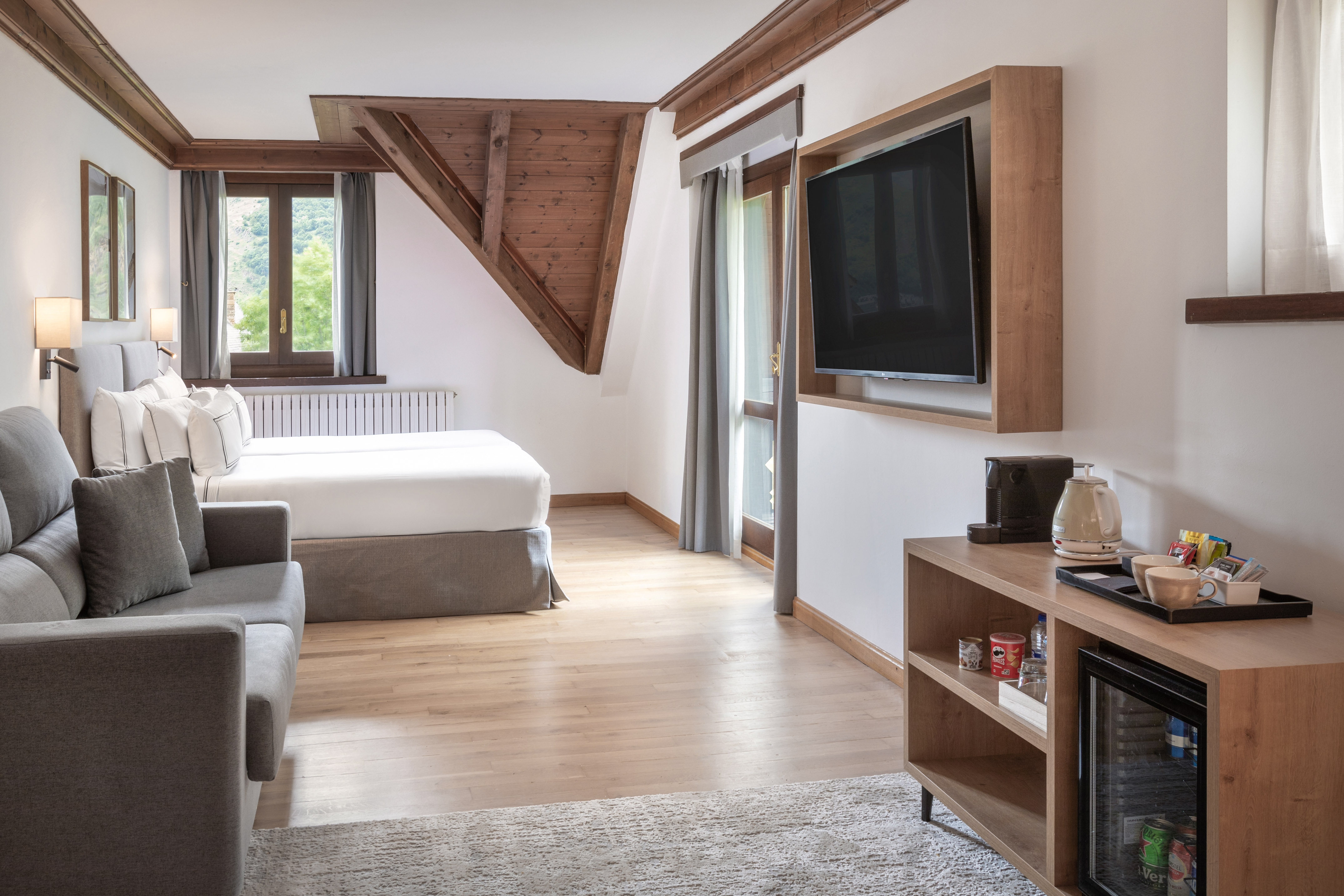 Modern hotel room with bed, sofa, TV, minibar, wooden beams, and scenic window.