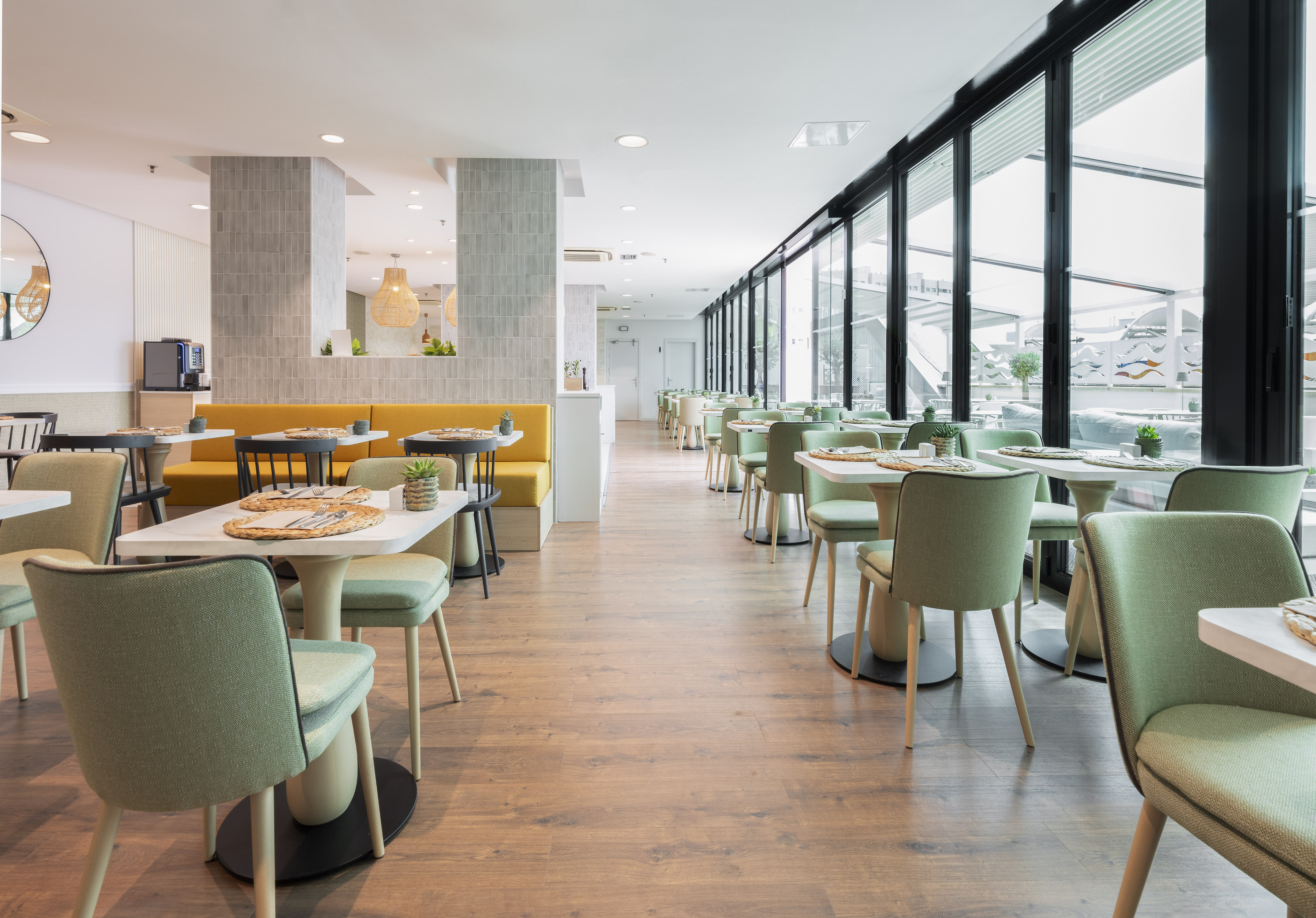 Modern restaurant dining room with light green chairs, yellow booths, and large windows.