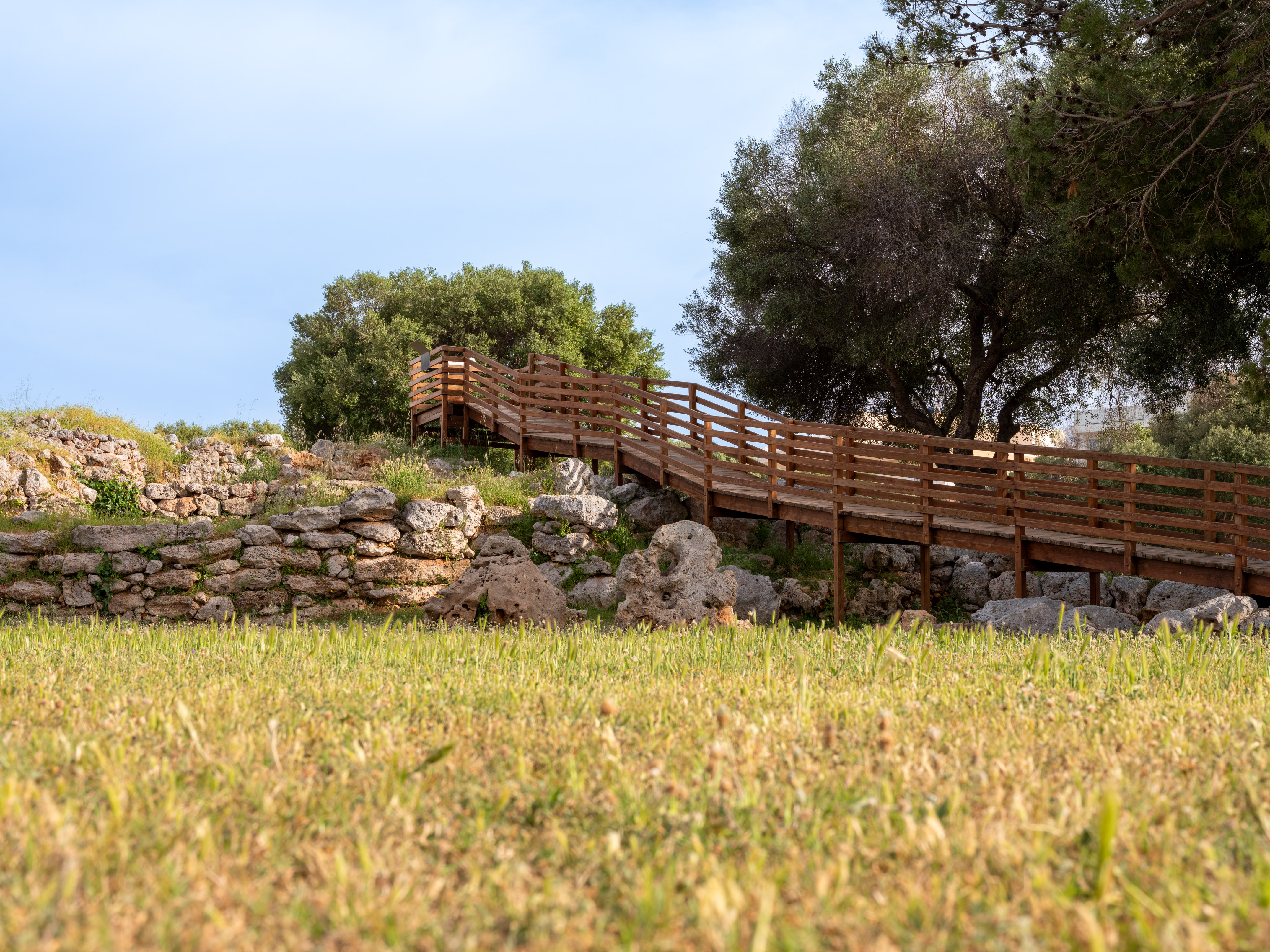 Wooden boardwalk ascending a rocky, grassy slope with trees and stone walls.