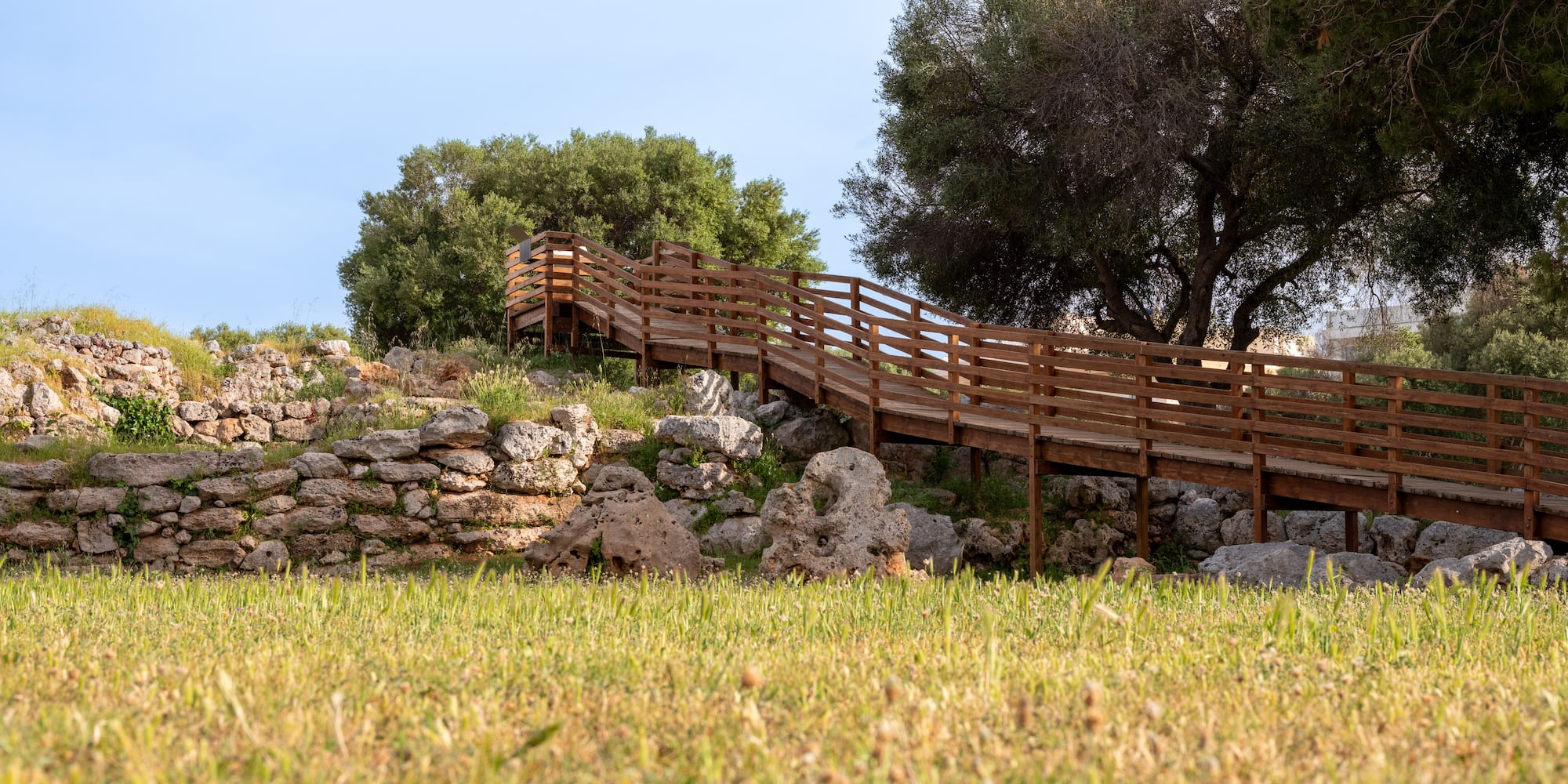 Wooden boardwalk ascending a rocky, grassy slope with trees and stone walls.