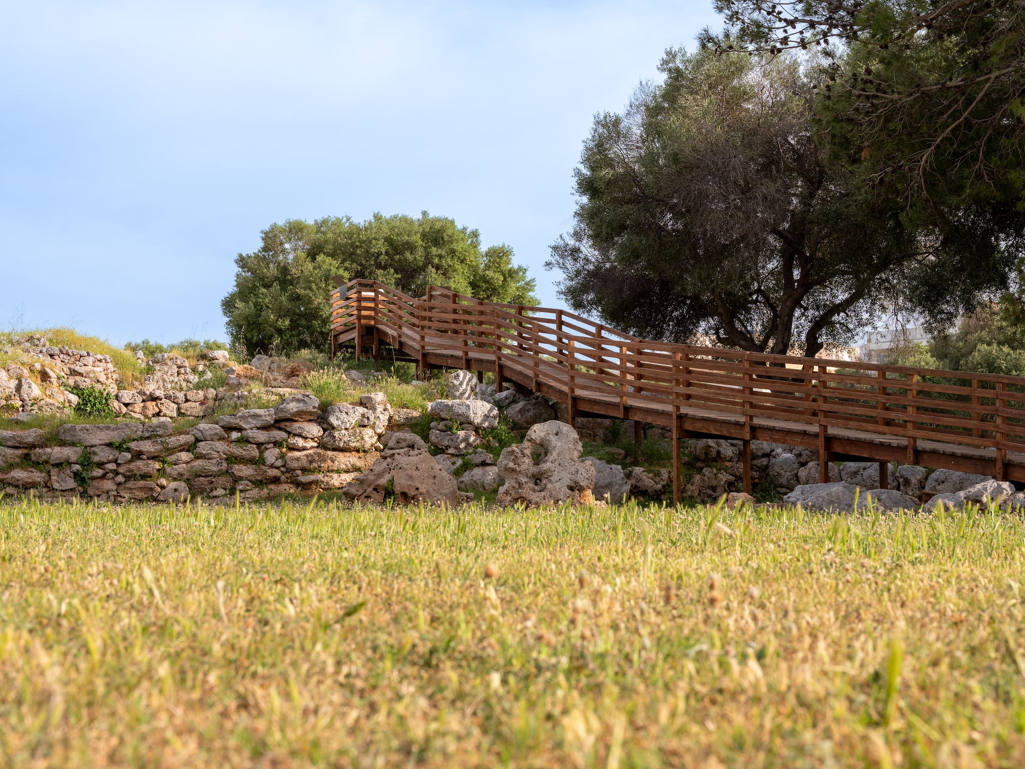 Wooden boardwalk ascending a rocky, grassy slope with trees and stone walls.