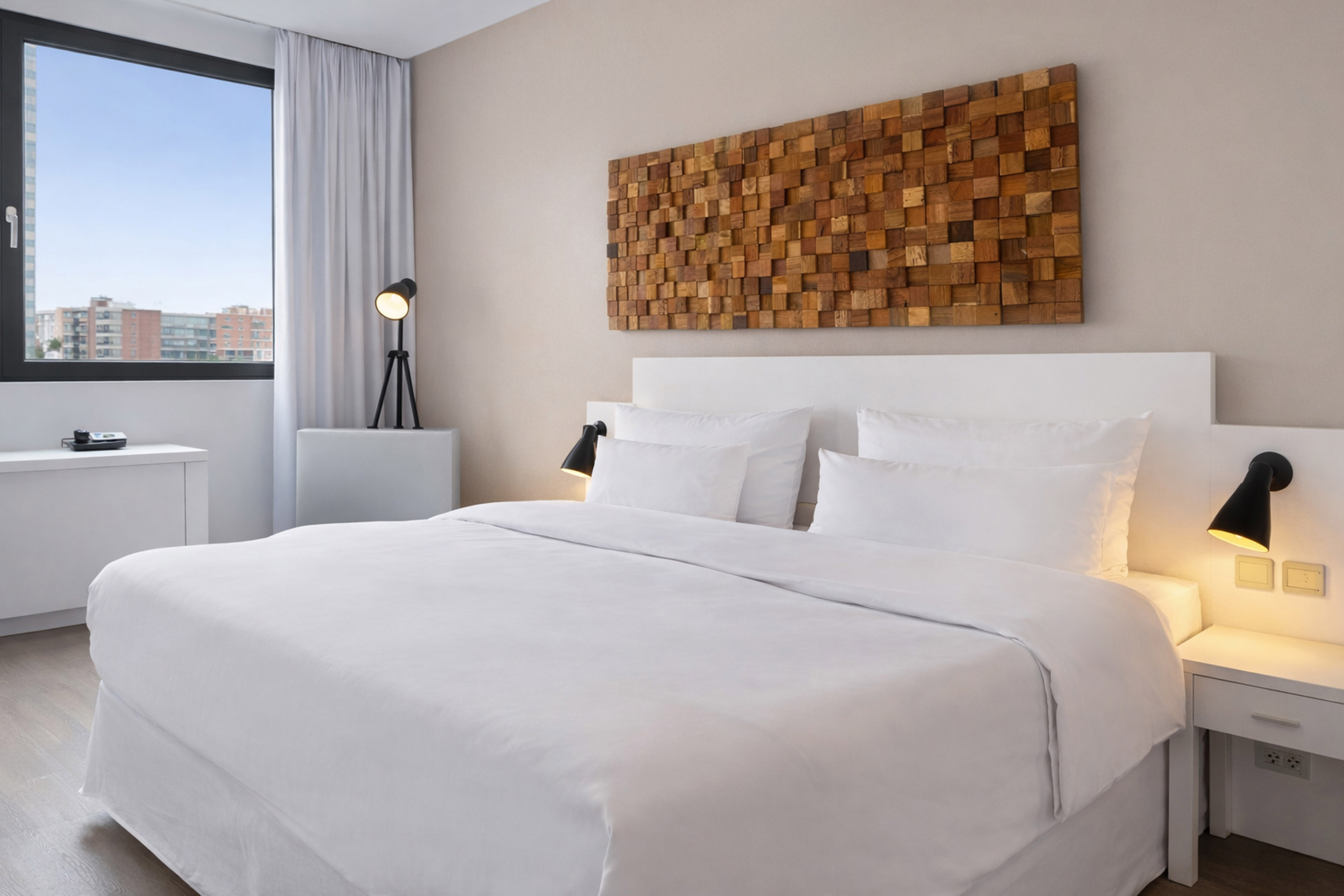 Modern hotel bedroom with white bed, wooden blocks art, and city view window.