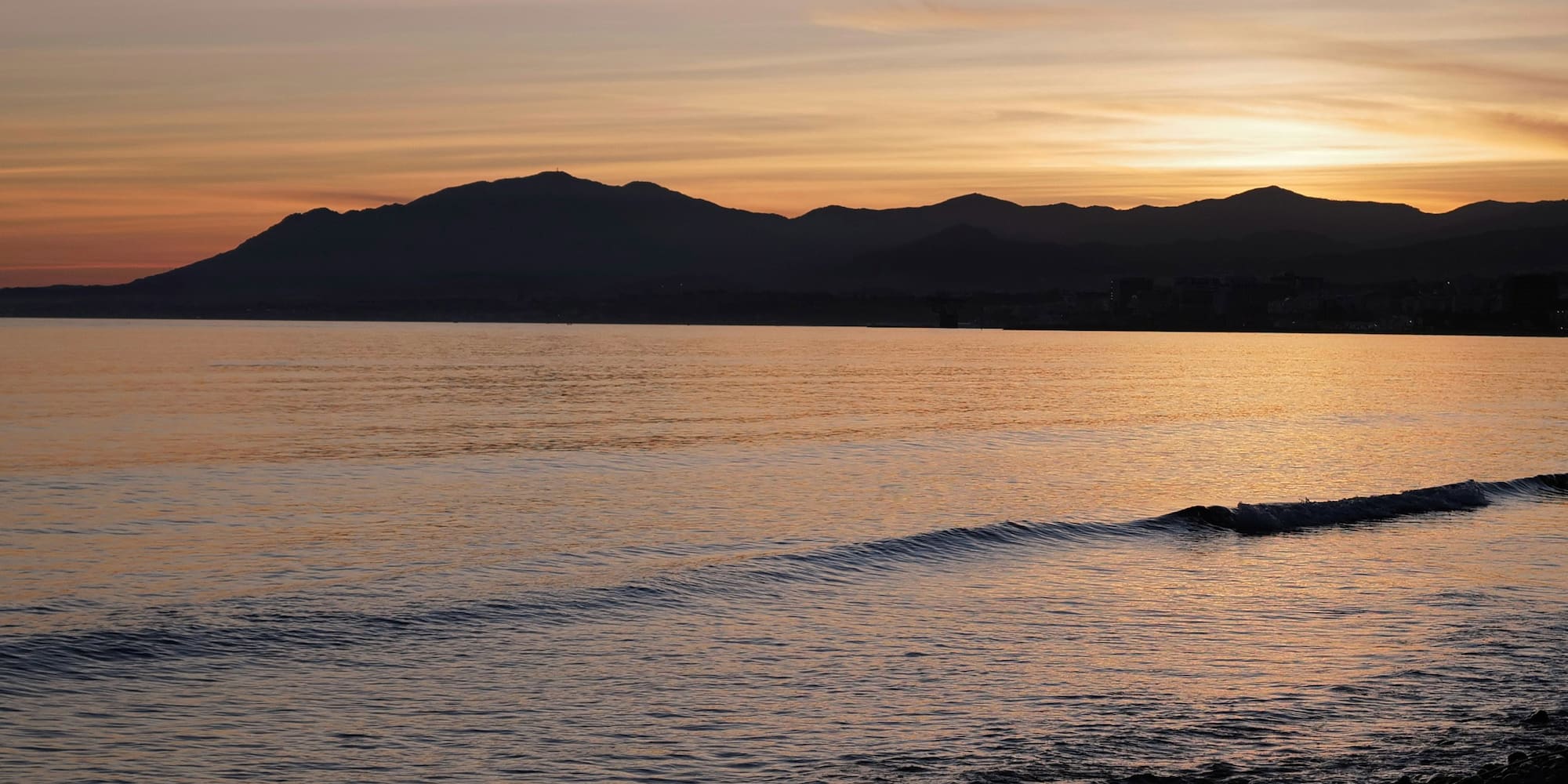 Golden sunset reflects on ocean waves, silhouetting coastal mountains.