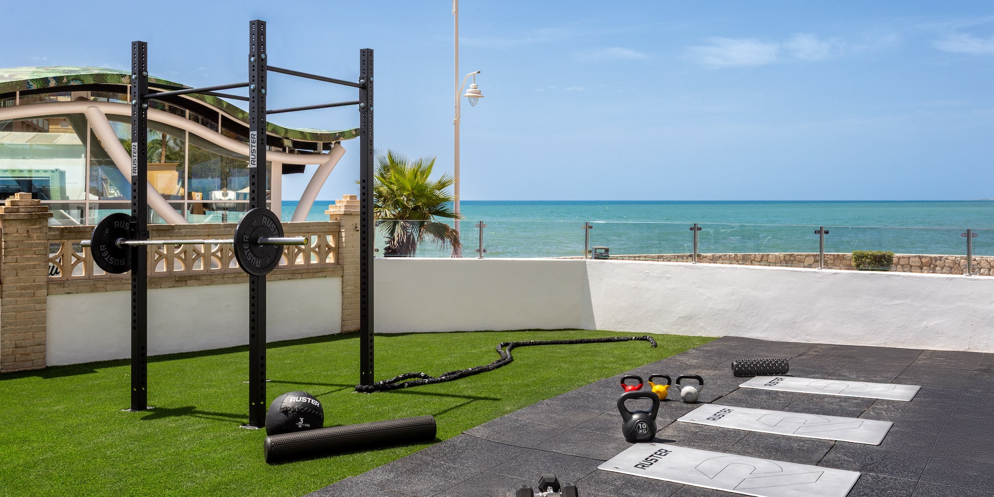 Beachfront outdoor gym with squat rack, kettlebells, dumbbells, and workout mats.