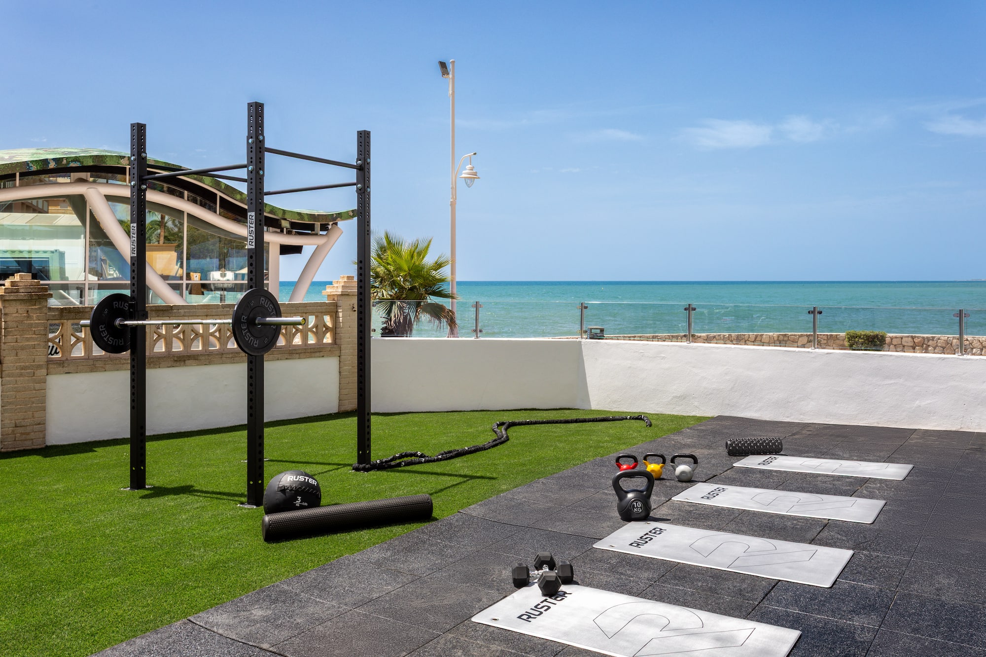 Outdoor gym overlooking ocean with squat rack, kettlebells, mats, and dumbbells.