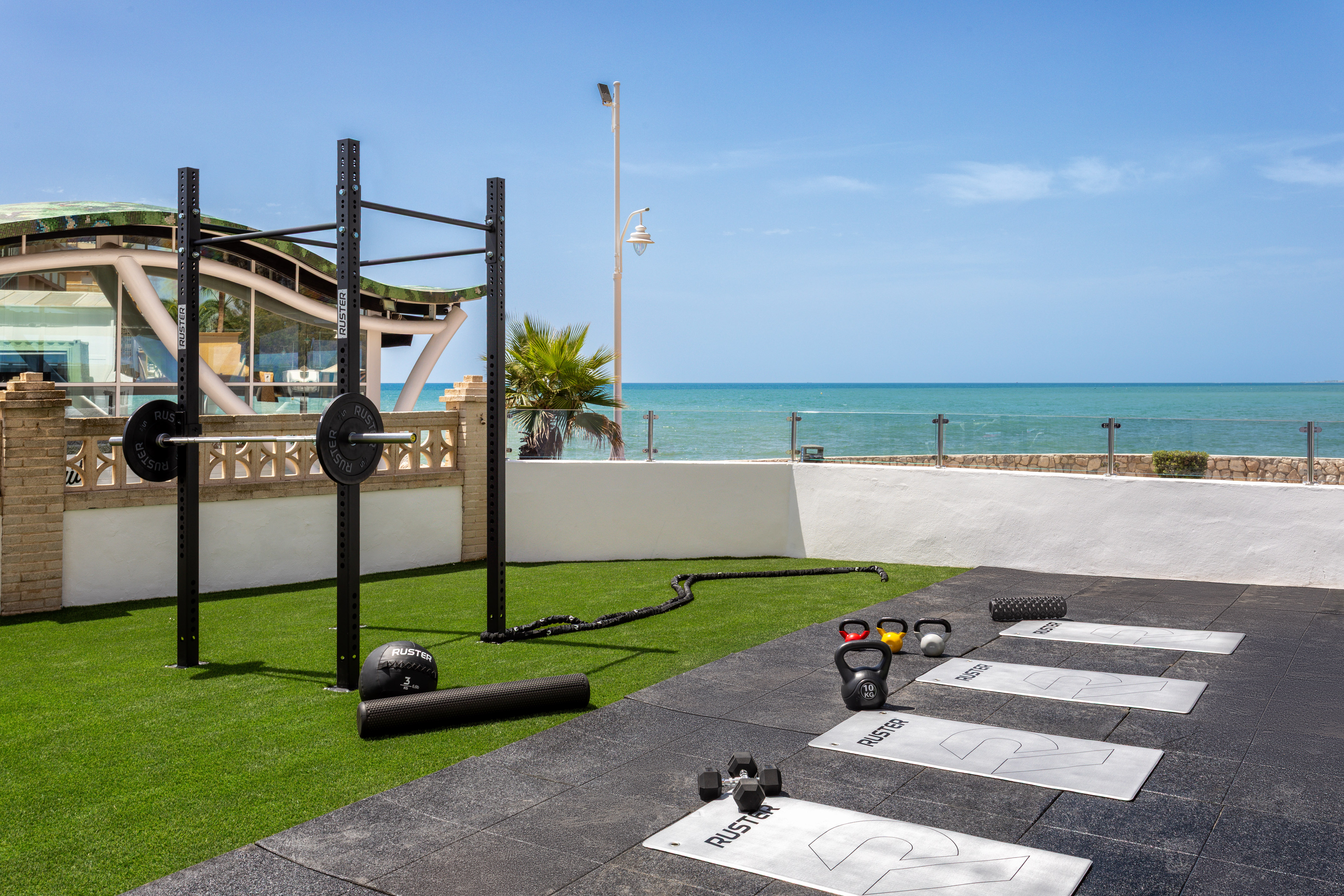 Outdoor gym overlooking ocean with squat rack, kettlebells, mats, and dumbbells.